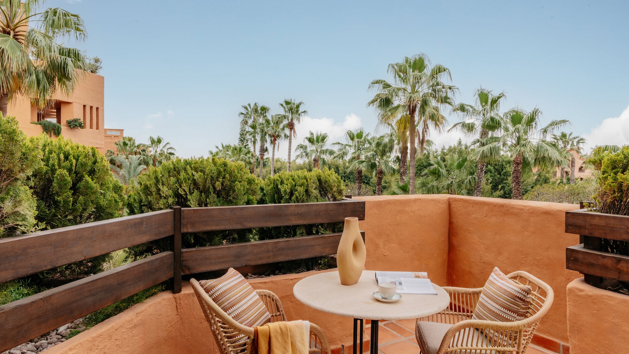 Terracotta balcony with wicker furniture, coffee, and tropical palm trees.