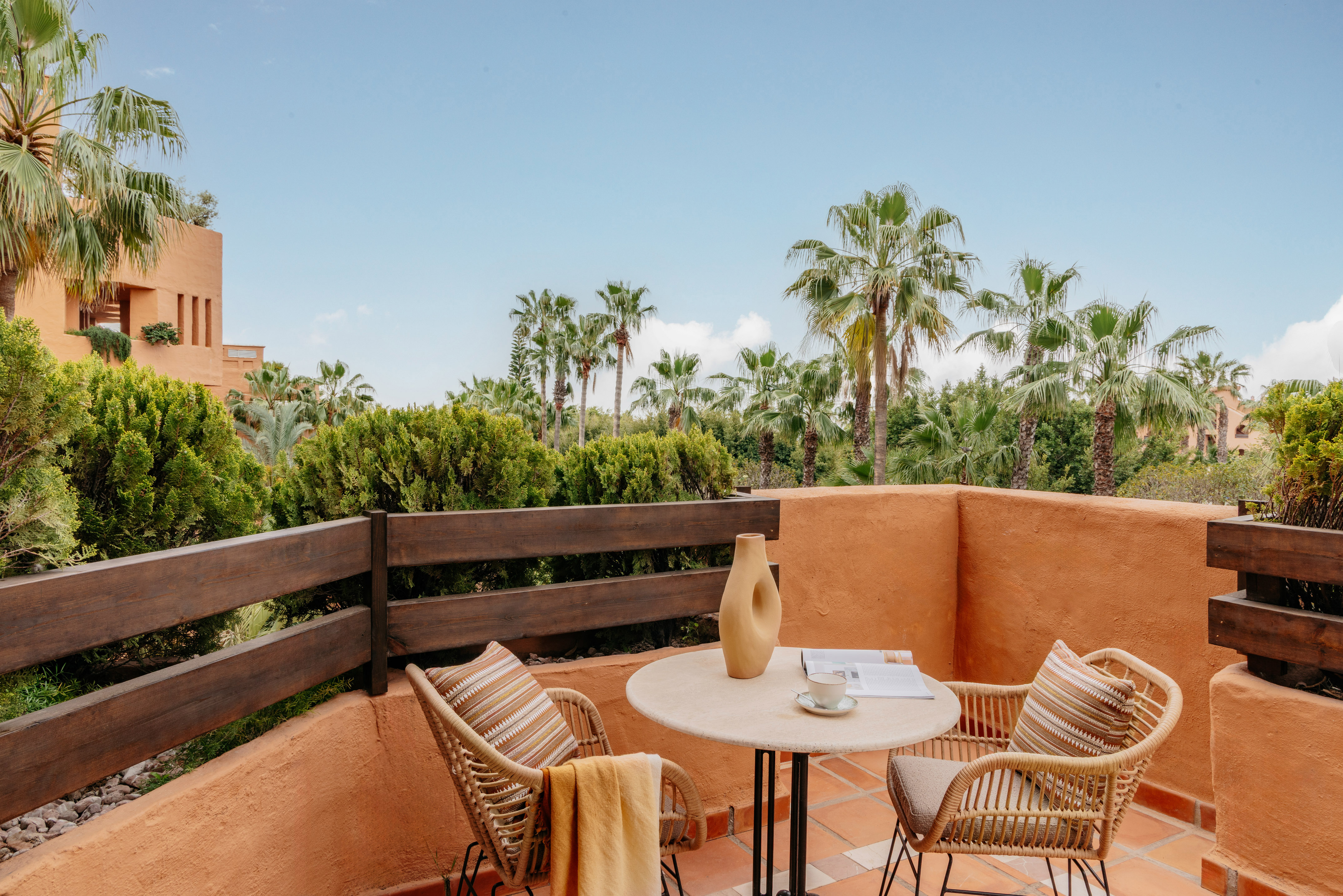 Terracotta balcony with wicker furniture, coffee, and tropical palm trees.