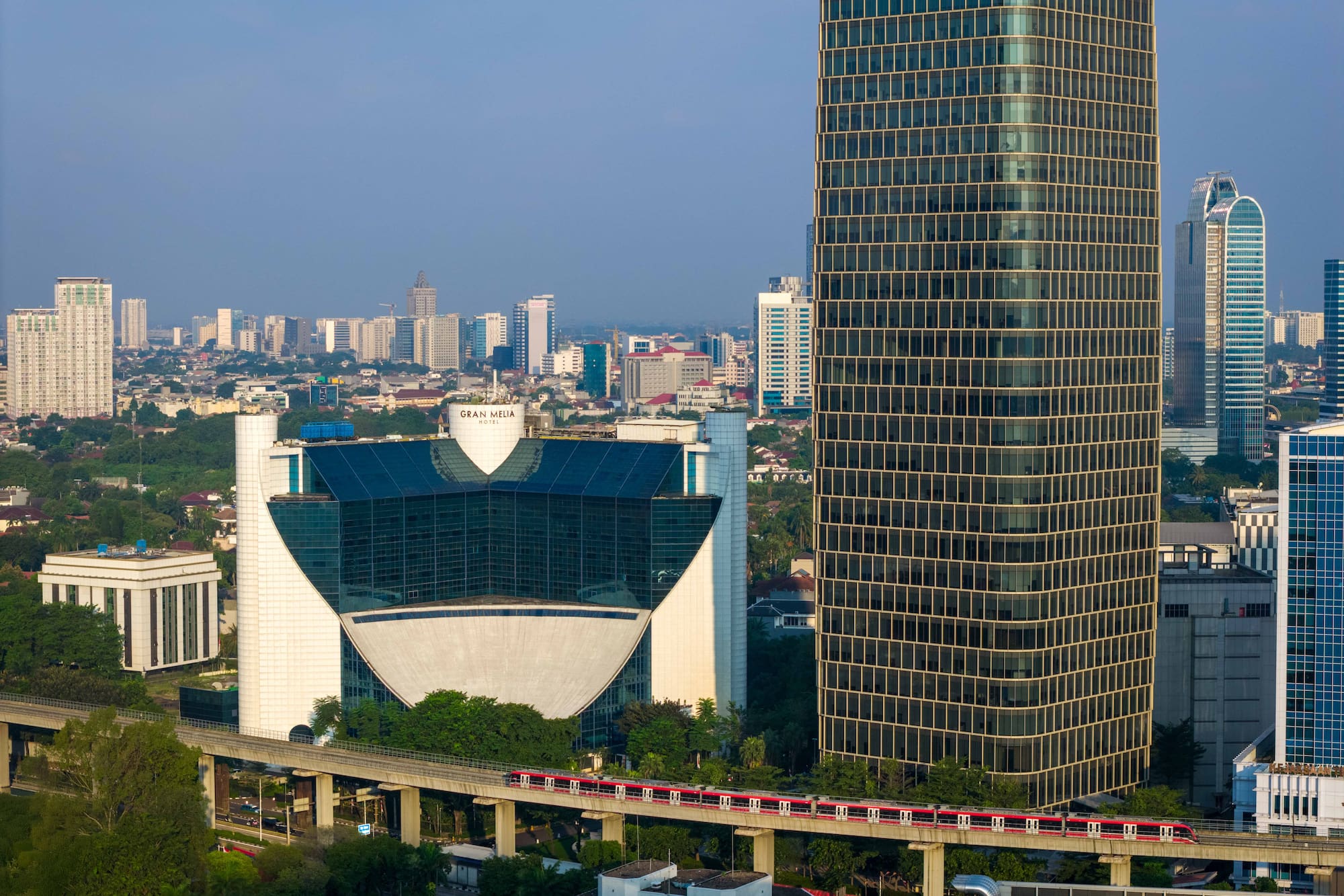 Jakarta skyline with modern buildings, red elevated train, and Gran Melia Hotel.