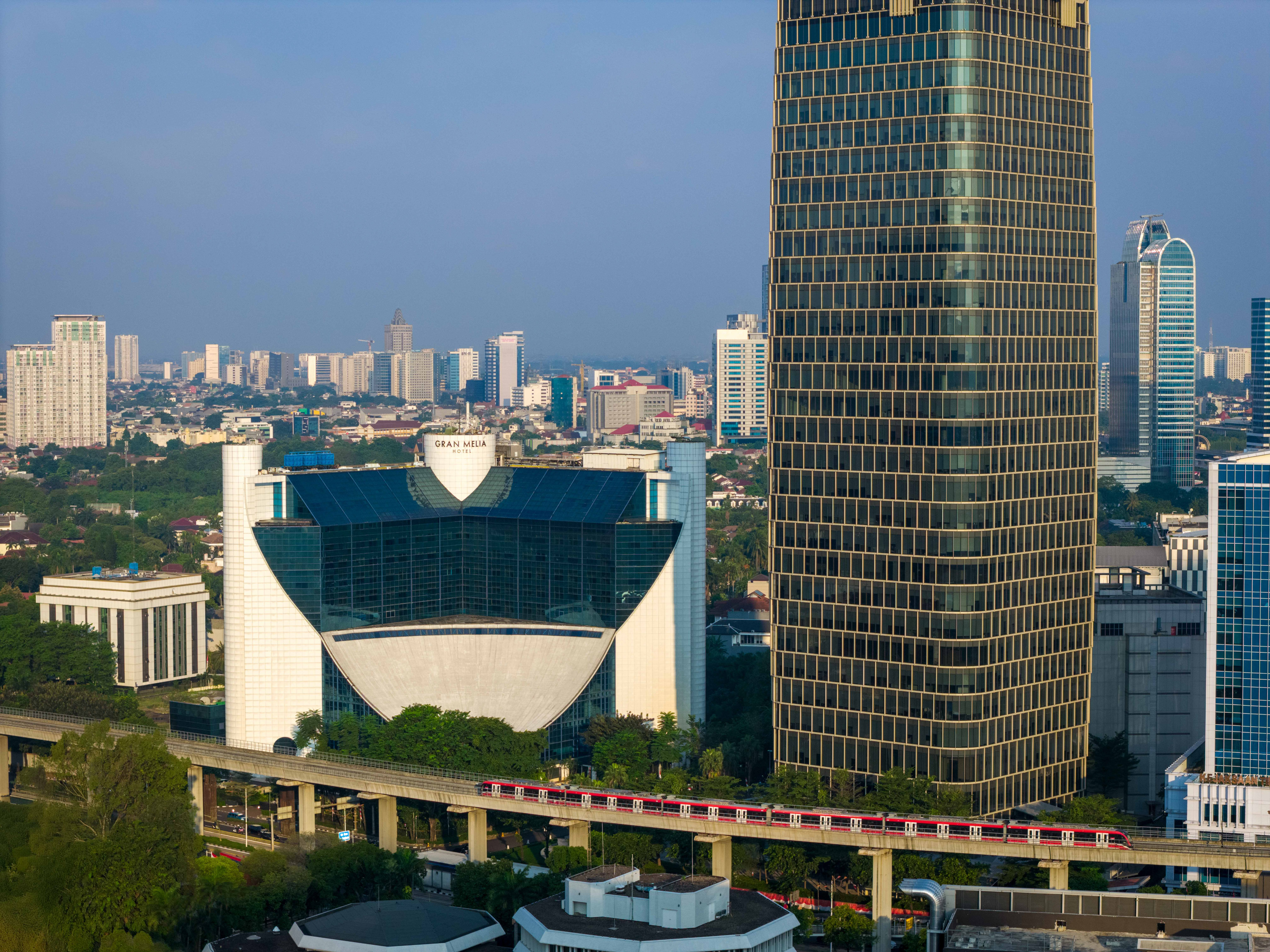 Jakarta skyline with modern buildings, red elevated train, and Gran Melia Hotel.