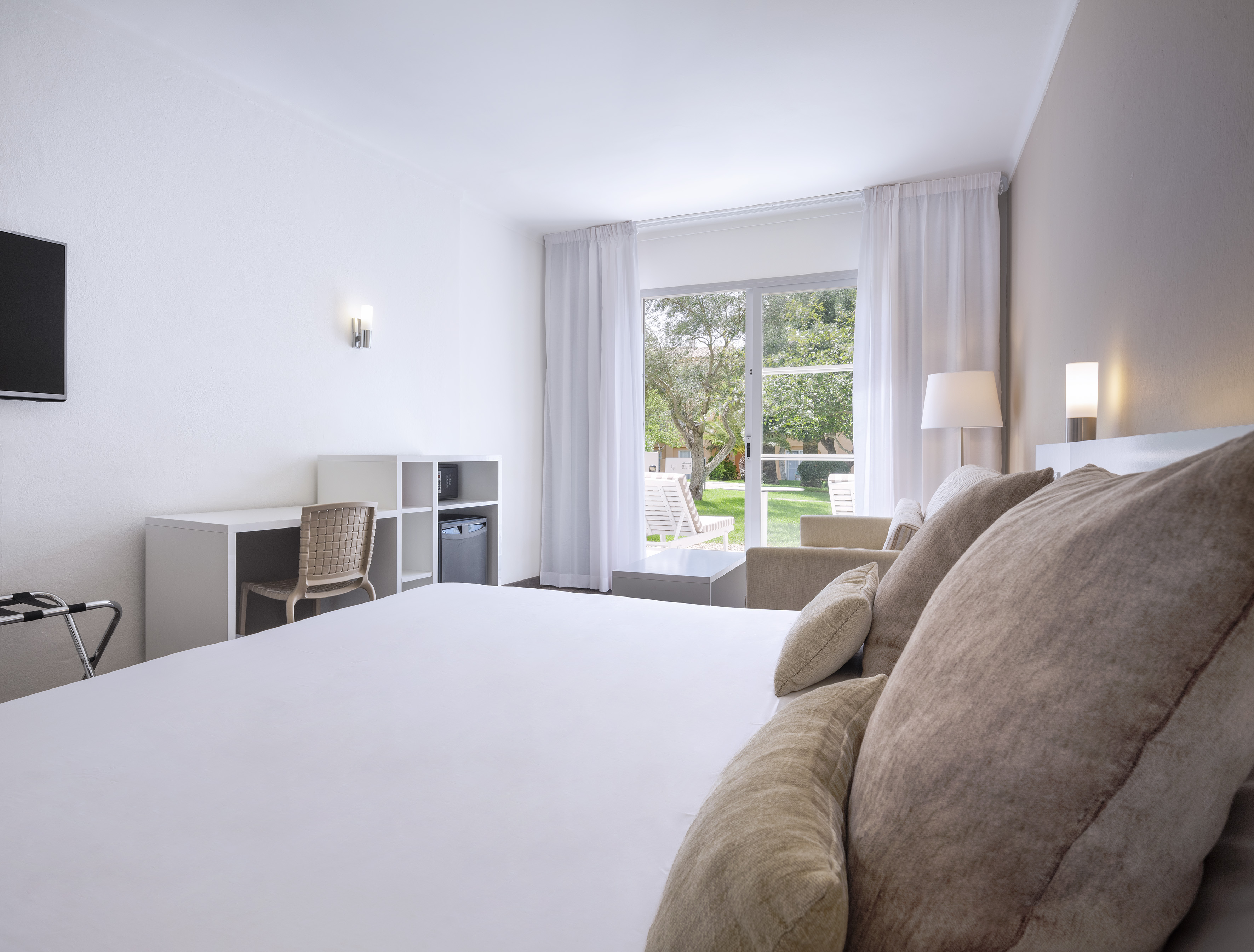 Modern hotel room with white bed, desk, minibar, and garden view patio.