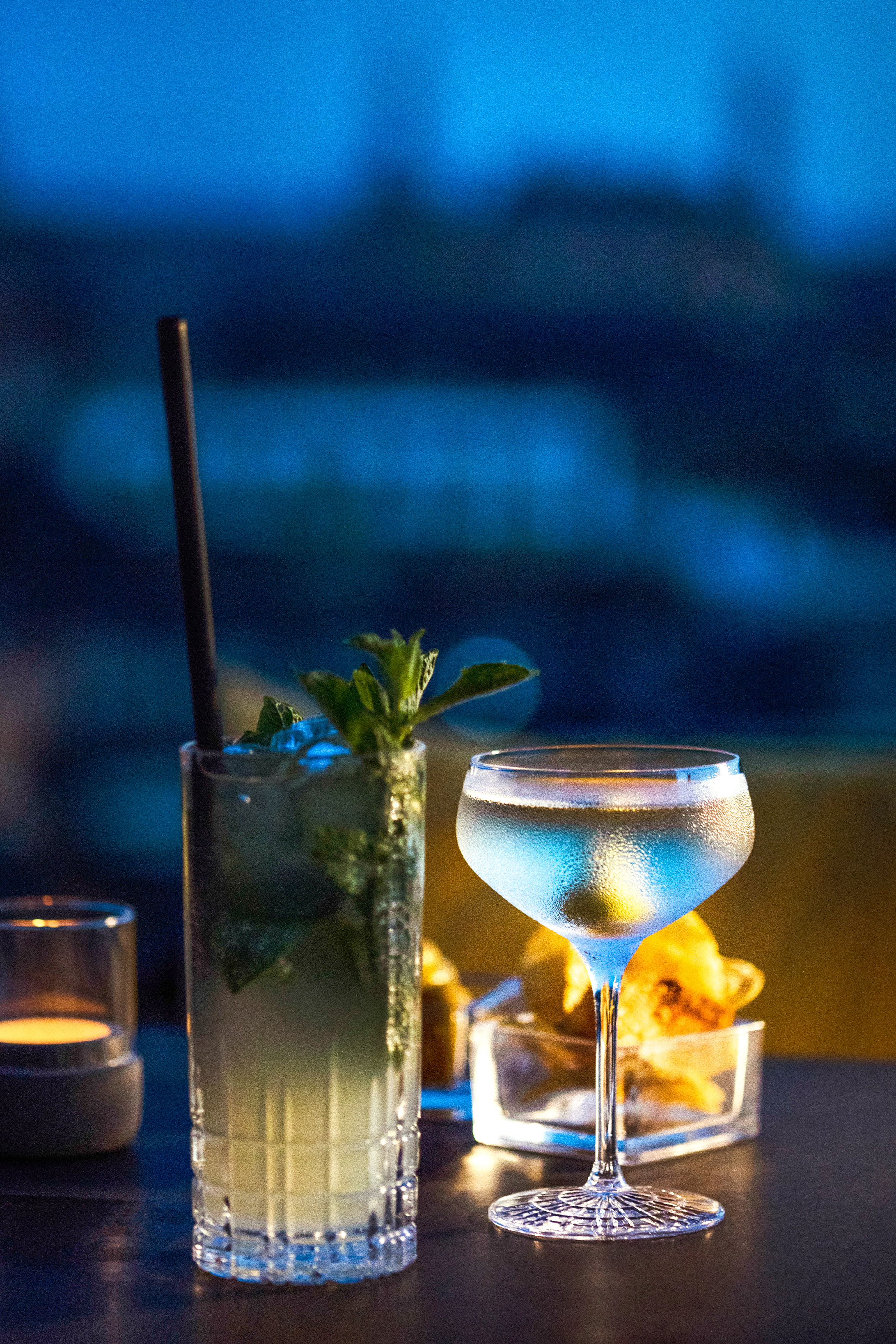 Mojito and martini cocktails with mint and olive on a bar table at night.
