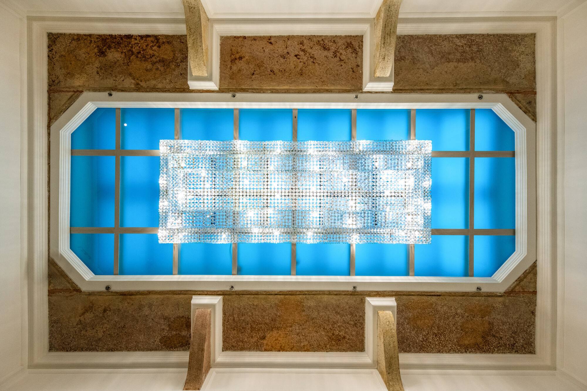 Modern ceiling with rectangular crystal chandelier over blue backlit panel.