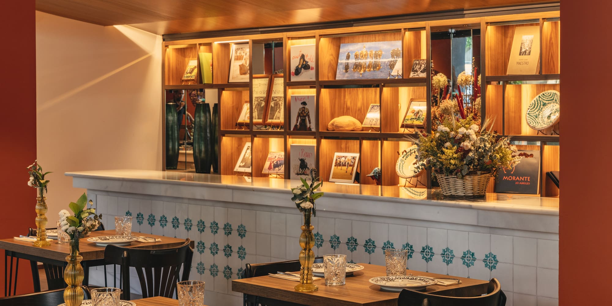 Restaurante español con mesas de madera, barra azulejada y estantería iluminada.