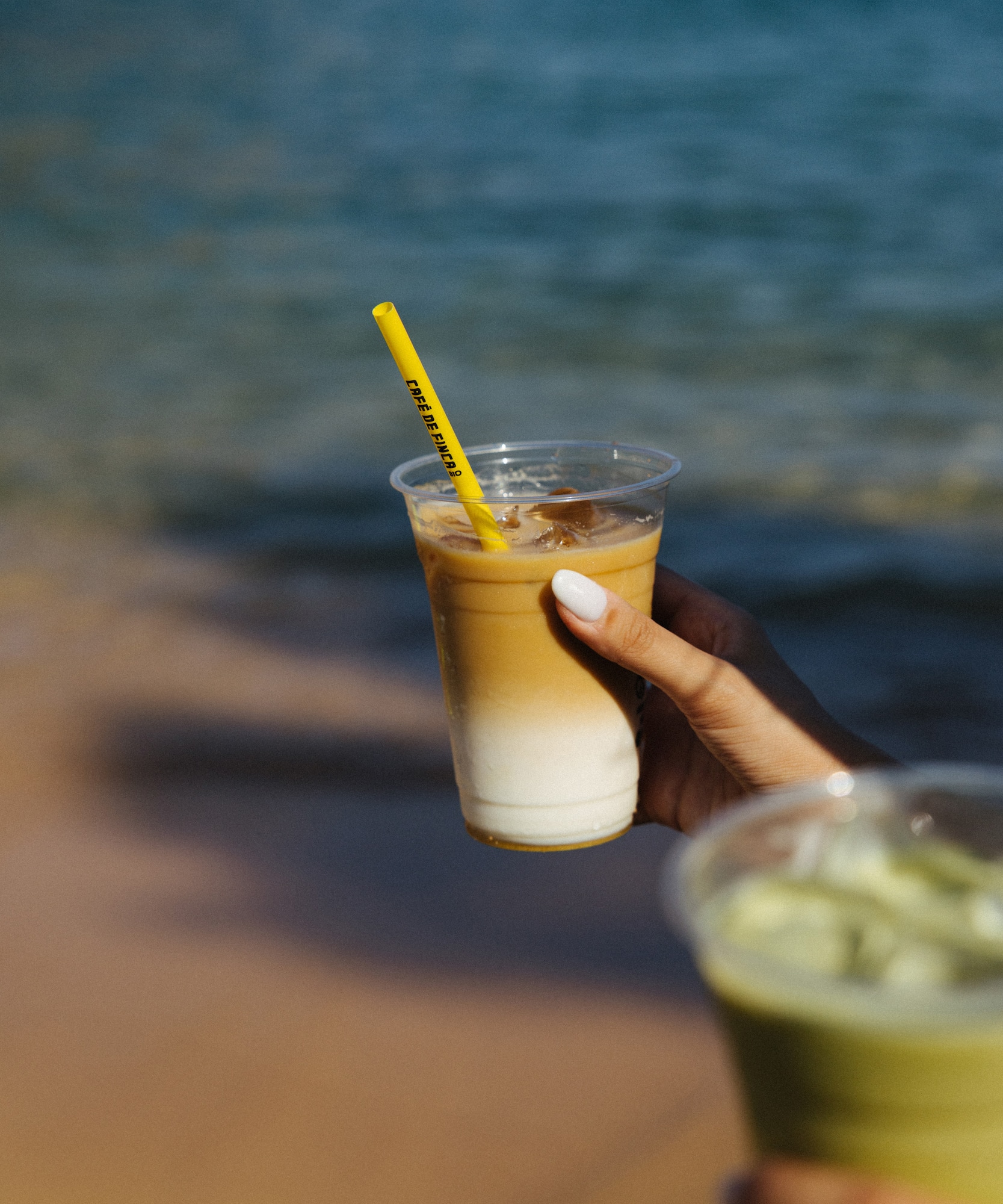 Hand holding layered iced latte with yellow 'CAFE DE FINCA' straw by the ocean.