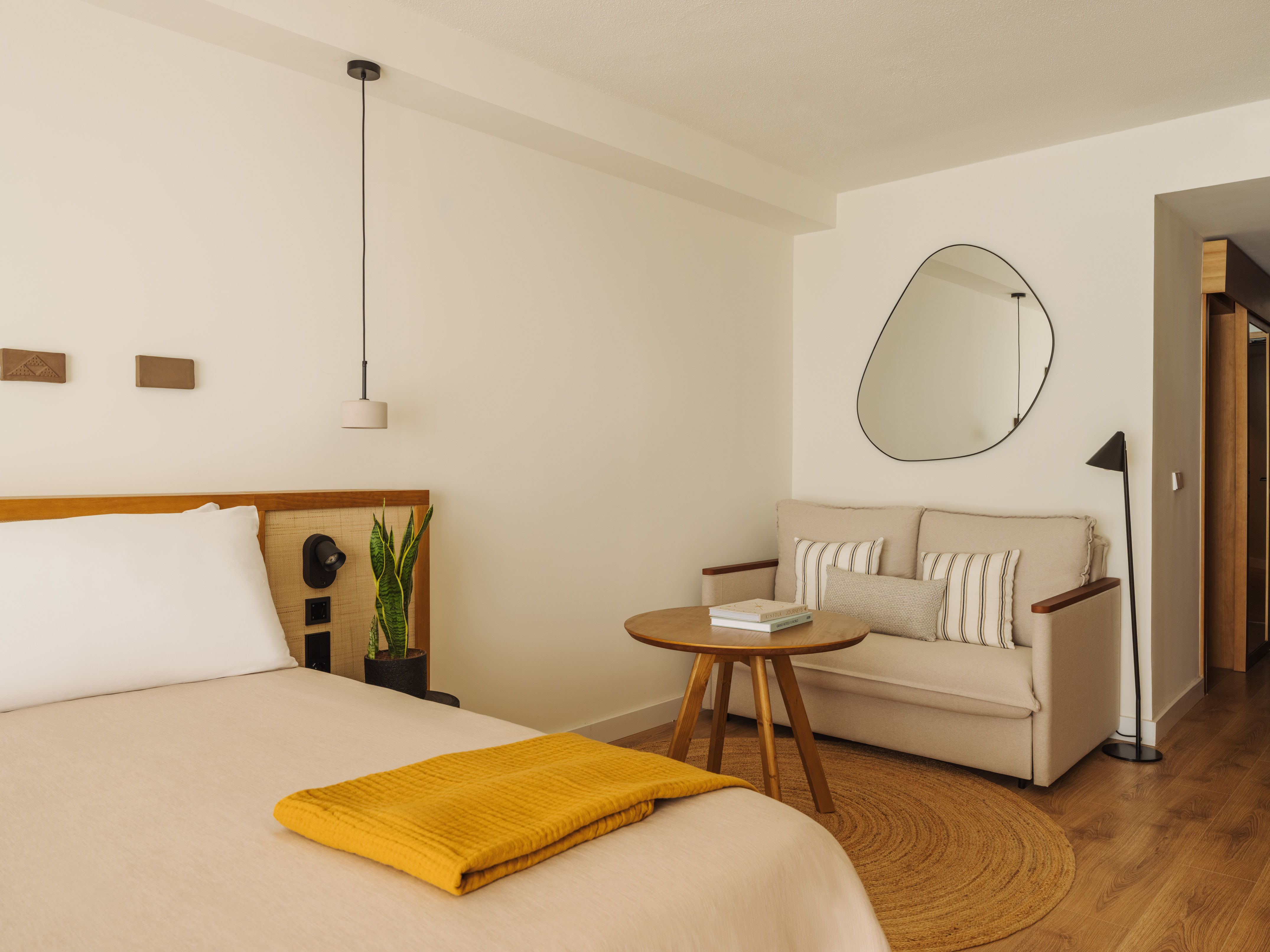 Stylish hotel room with beige bed, yellow blanket, sofa, and organic mirror.
