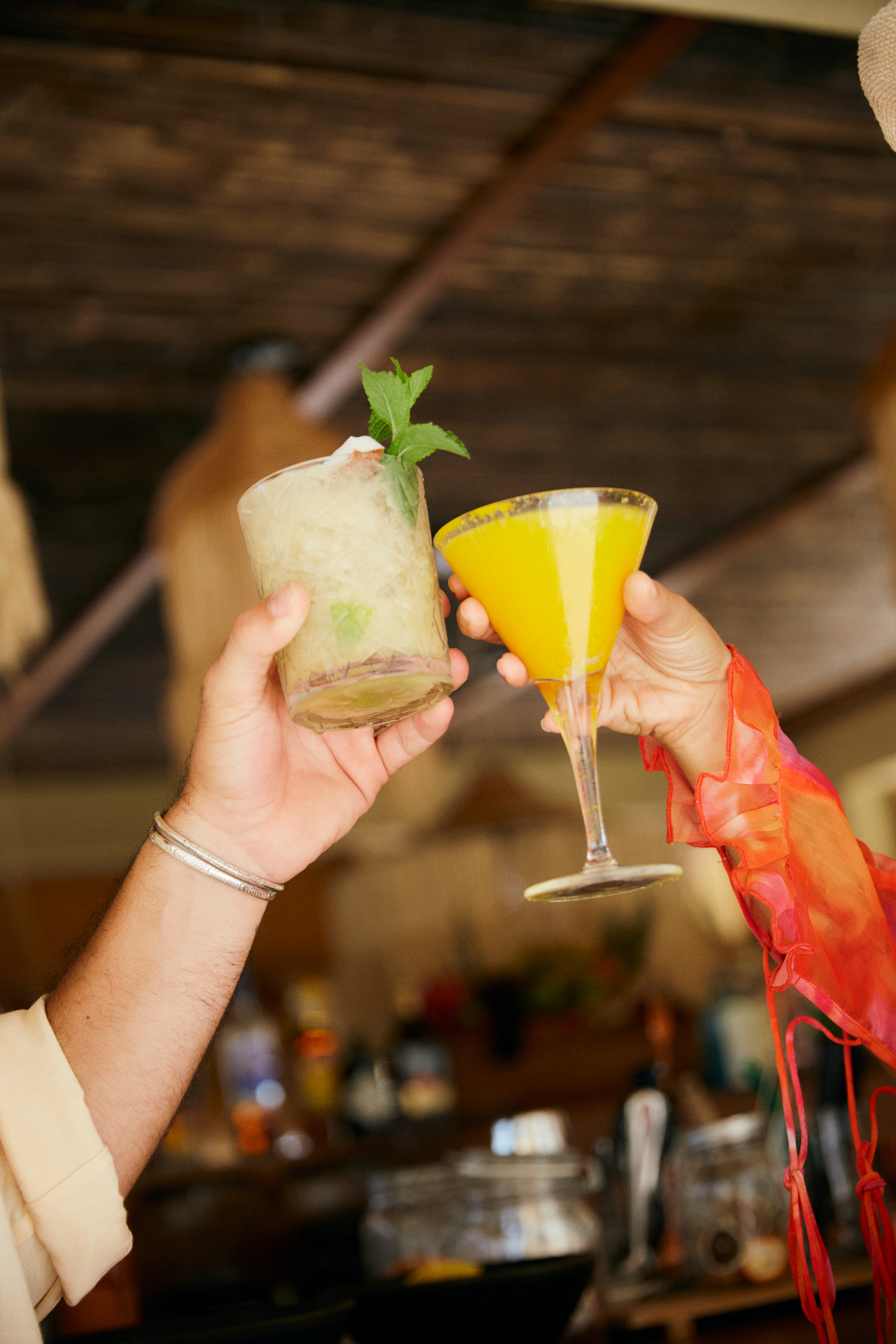 Hands clinking cocktail glasses: a mojito and a yellow martini at a bar.