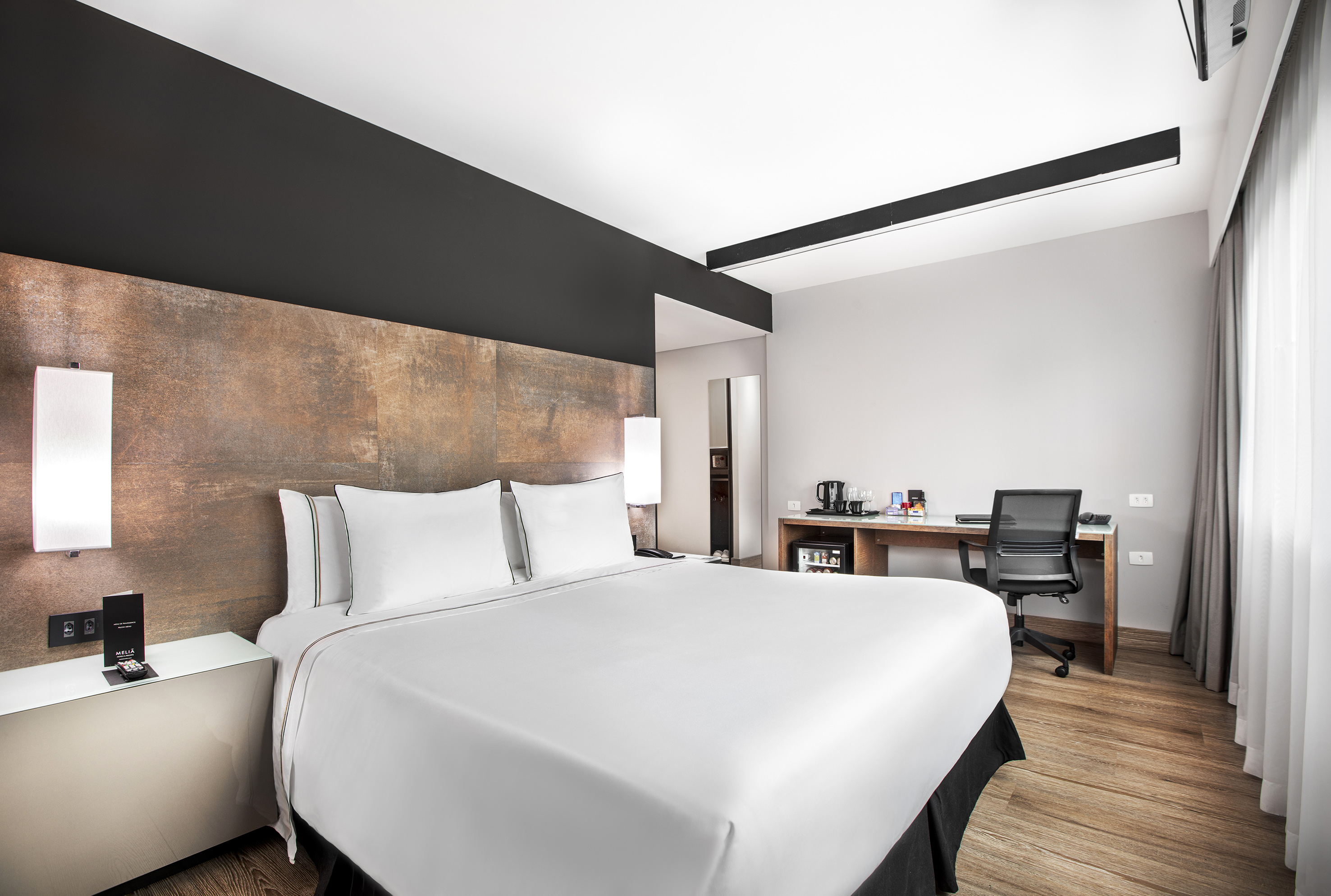 Modern hotel bedroom with white bed, textured headboard, and wood desk.