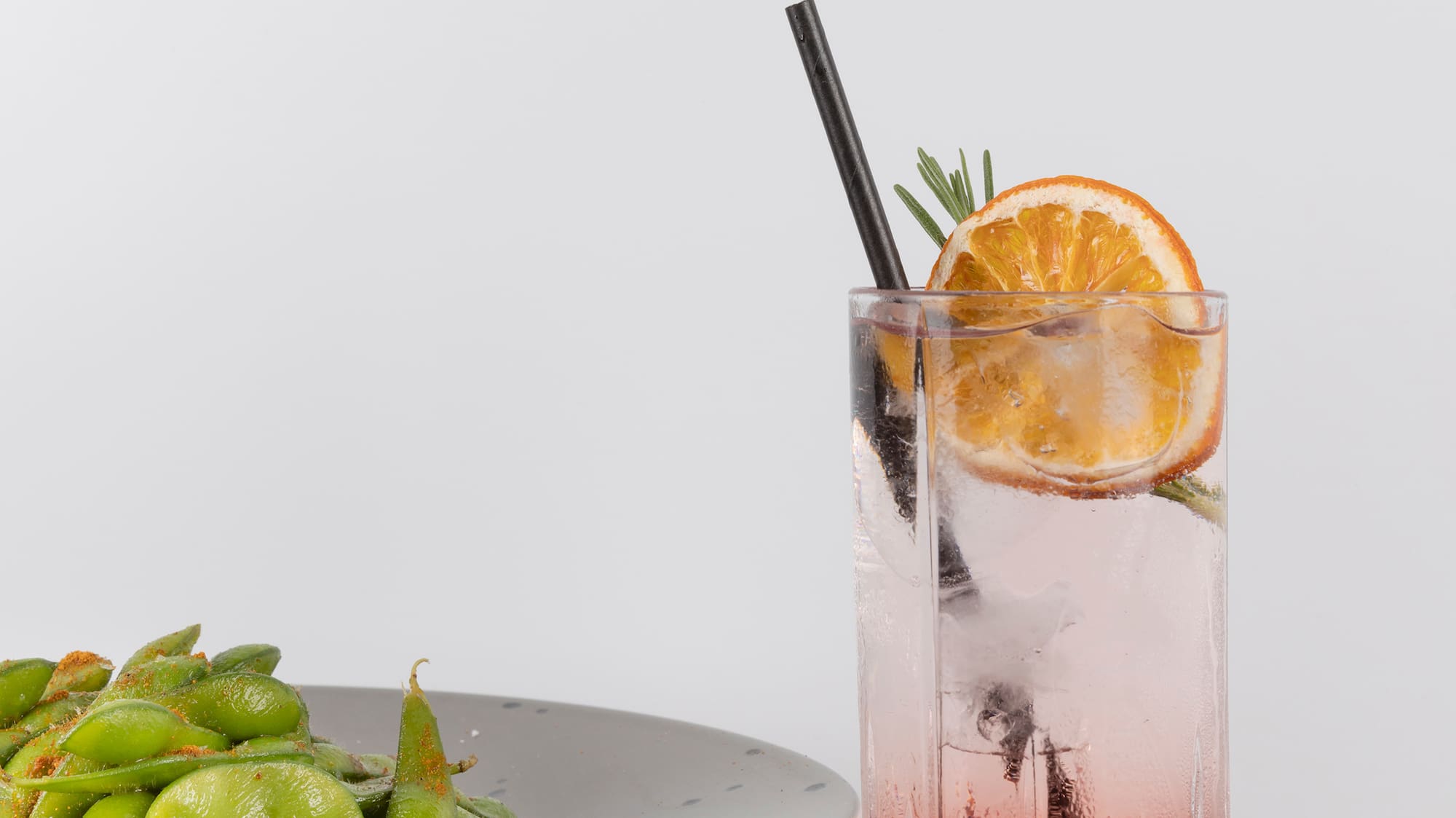 a glass of liquid with a slice of orange and a plate of edamame