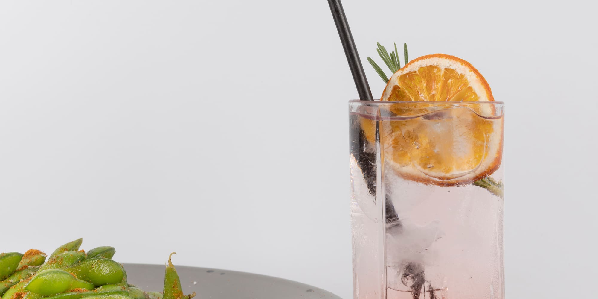 a glass of liquid with a slice of orange and a plate of edamame