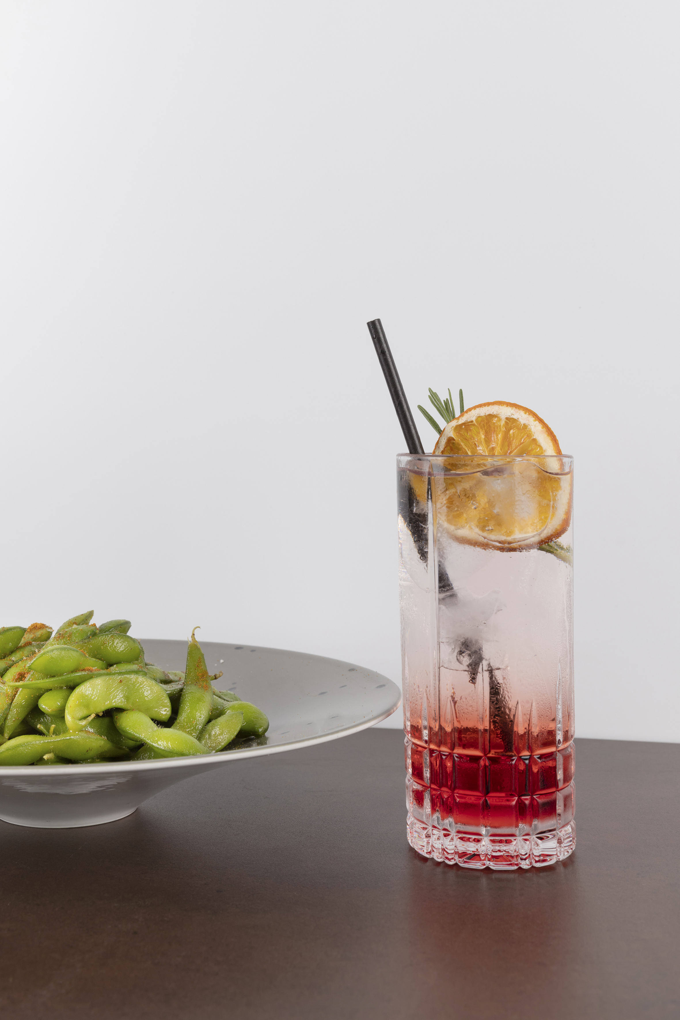 a glass of liquid with a slice of orange and a plate of edamame