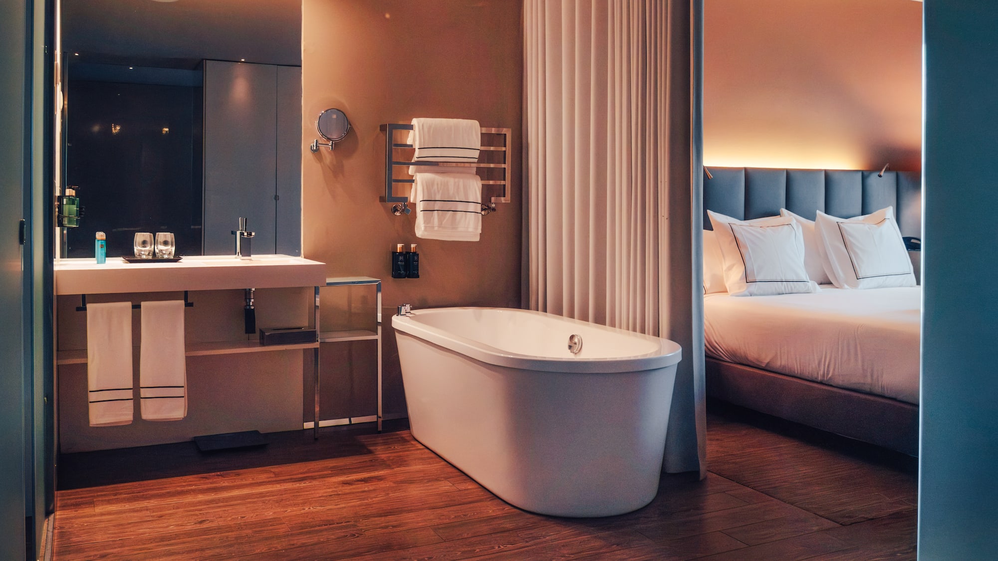 Modern hotel room with freestanding tub, wood floor, and bedroom.