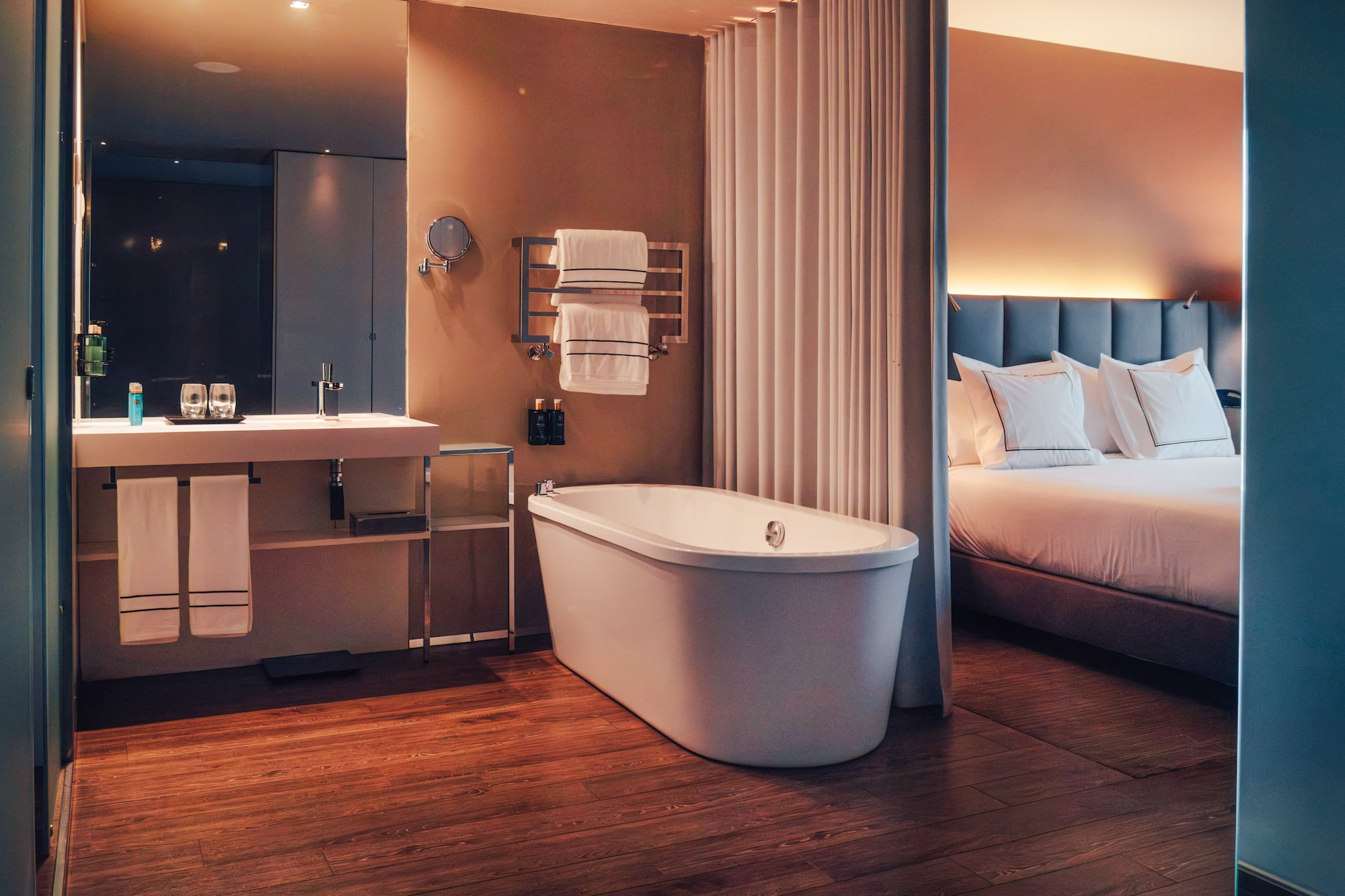 Modern hotel room with freestanding tub, wood floor, and bedroom.
