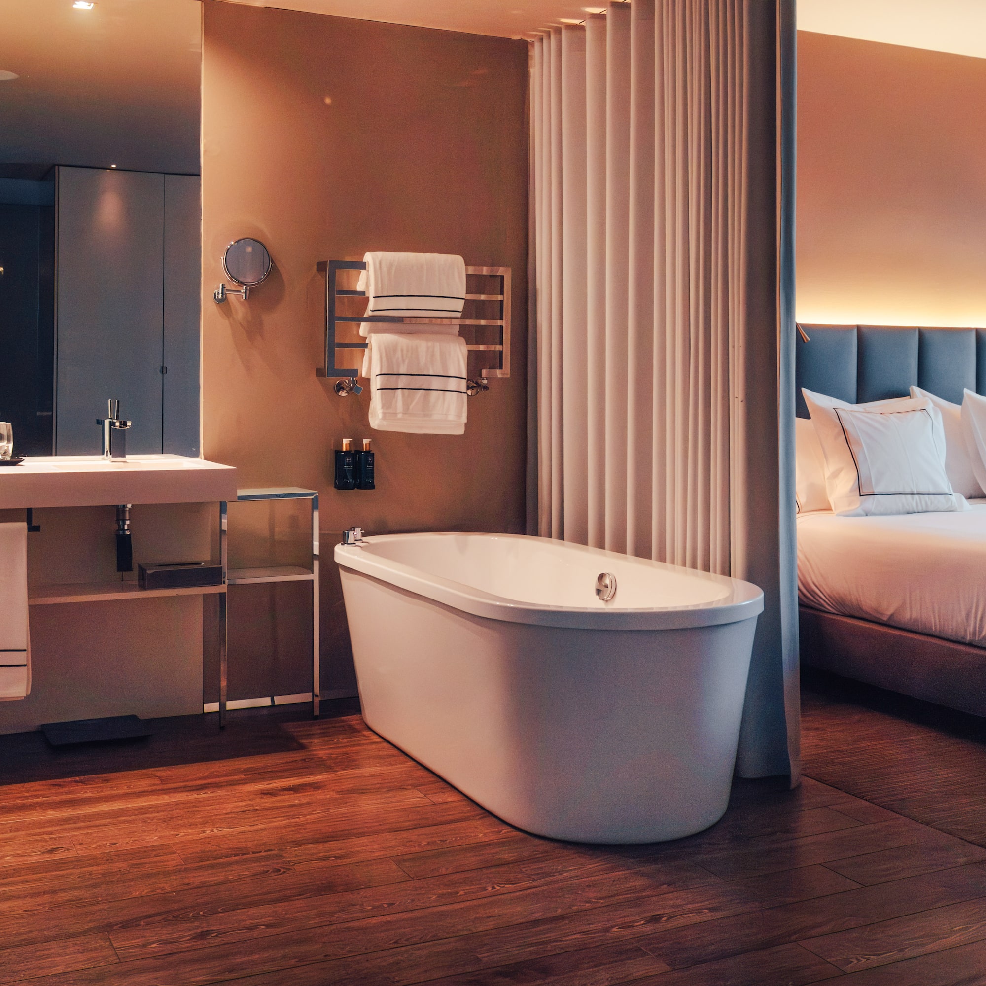 Modern hotel room with freestanding tub, wood floor, and bedroom.