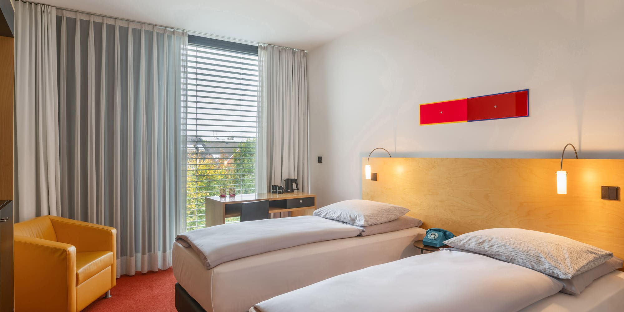 Modern hotel room with twin beds, red carpet, orange chair, and desk.