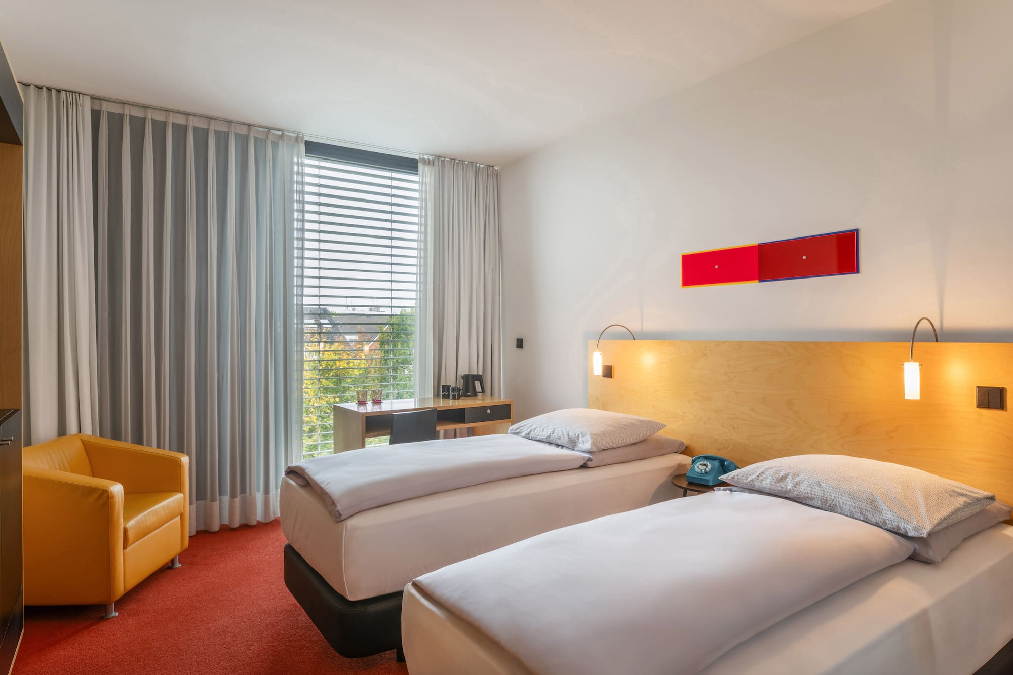 Modern hotel room with twin beds, red carpet, orange chair, and desk.
