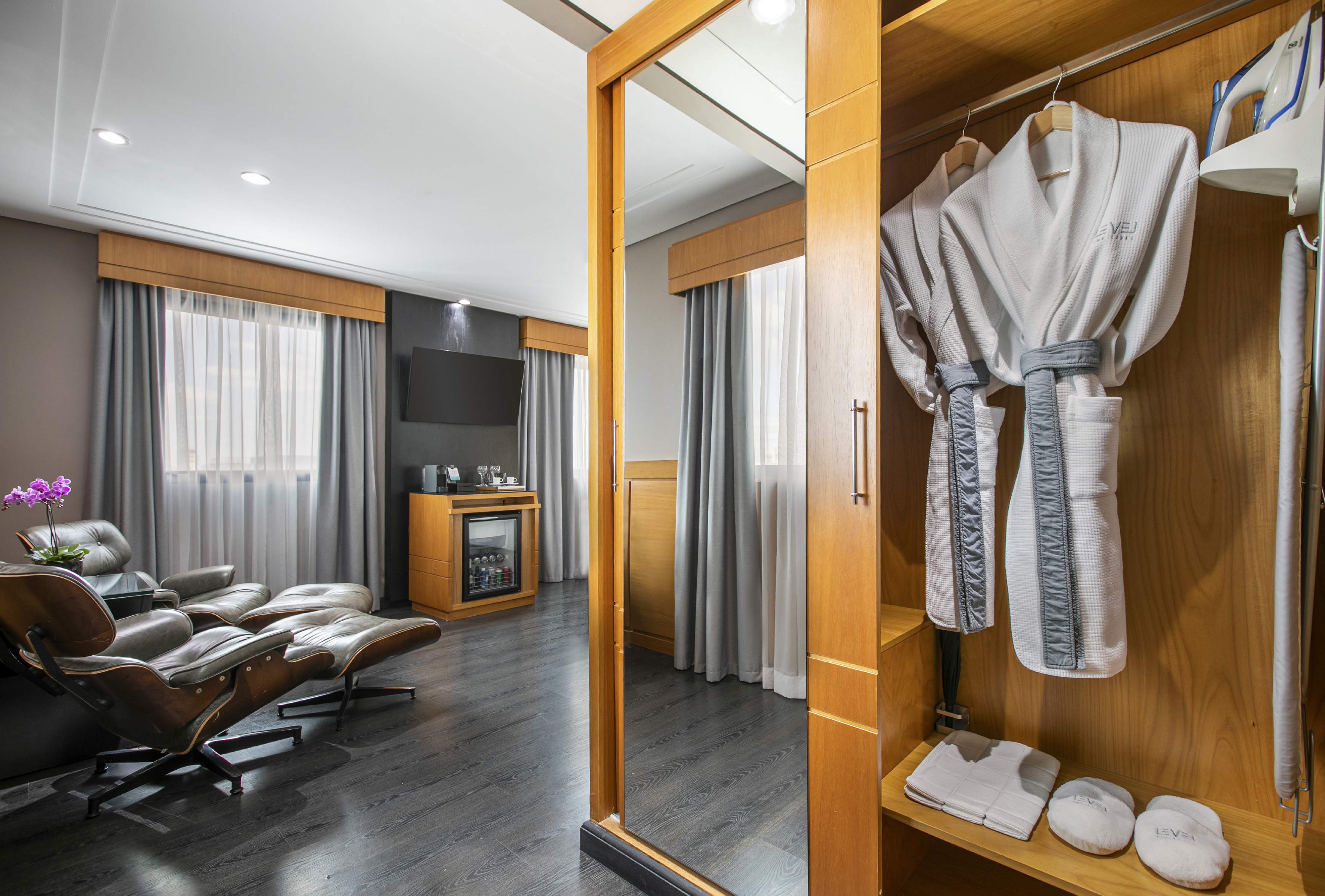 Hotel room wardrobe with bathrobes, slippers, mirror, lounge chairs, and minibar.
