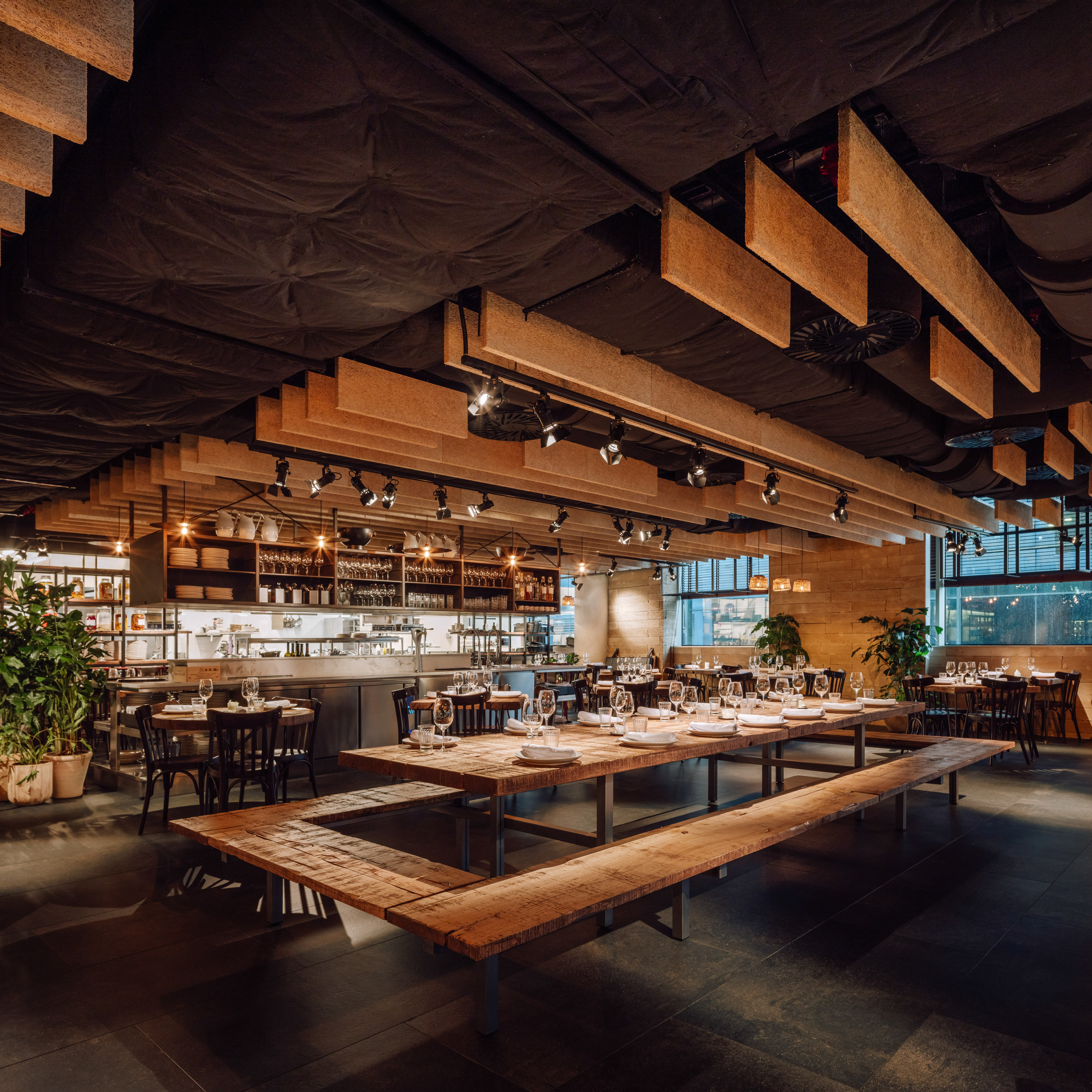 Restaurant interior with wooden communal tables, benches, and open kitchen.