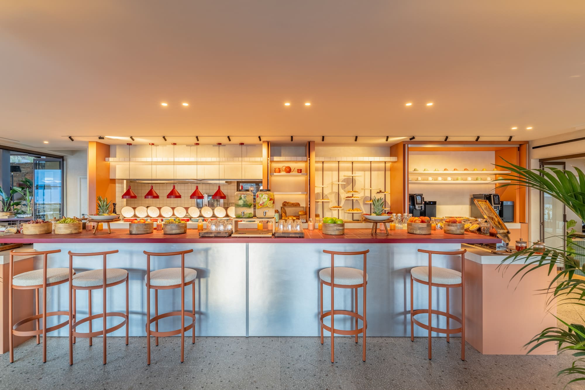 Modern hotel breakfast buffet with fresh fruit, juices, and bar stools.
