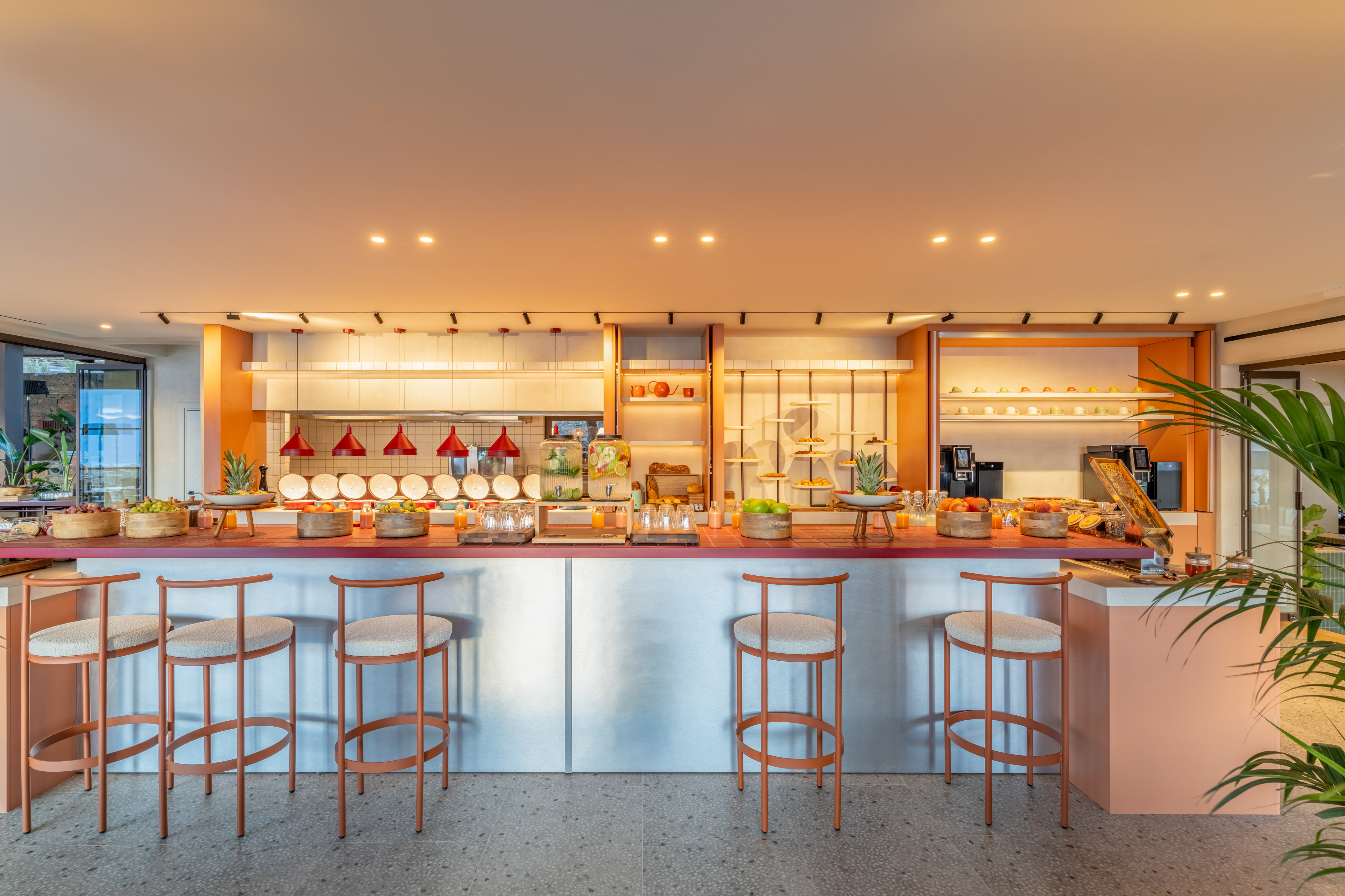 Modern hotel breakfast buffet with fresh fruit, juices, and bar stools.