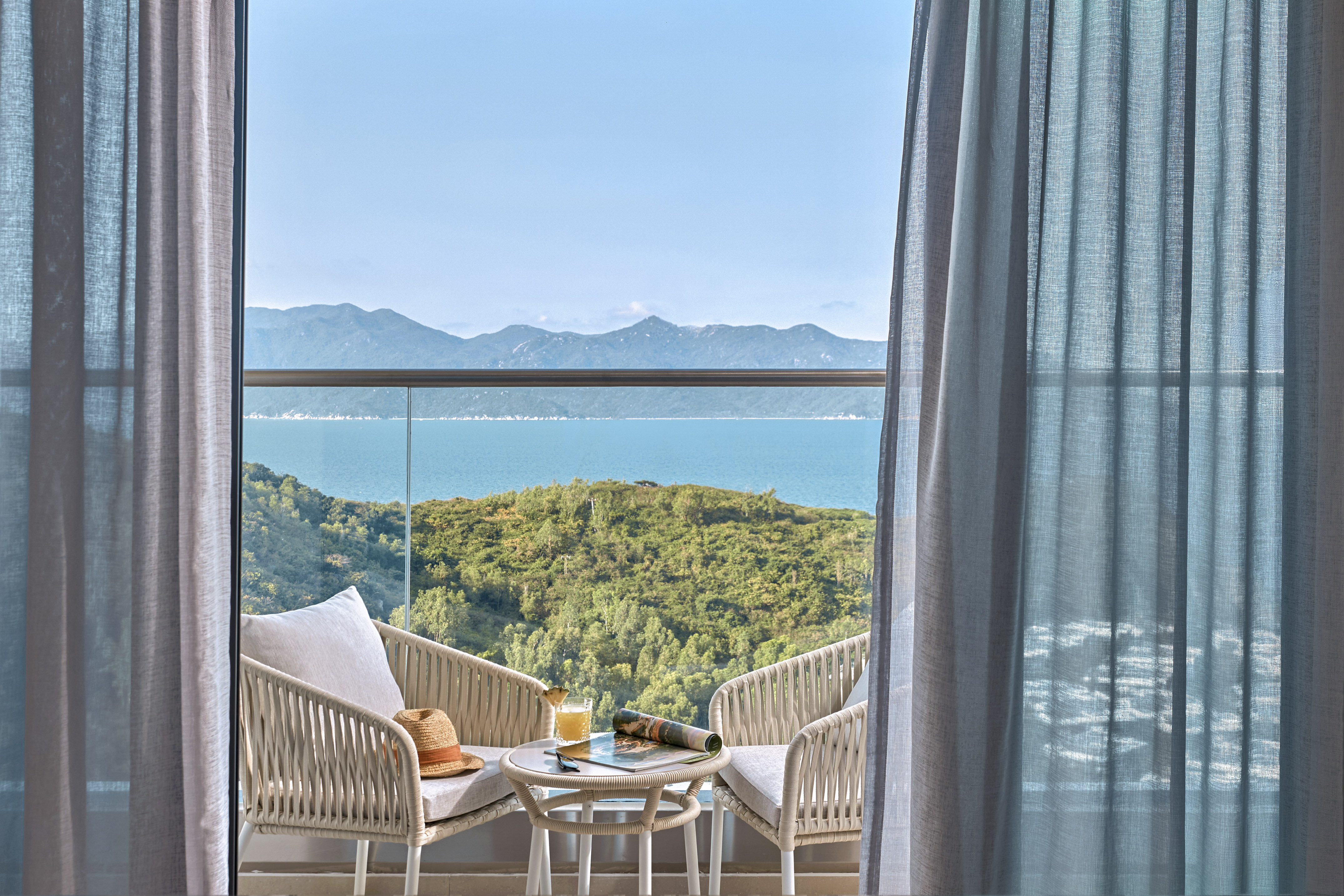 Balcony with sea view, outdoor seating, green hills, and blue sky.