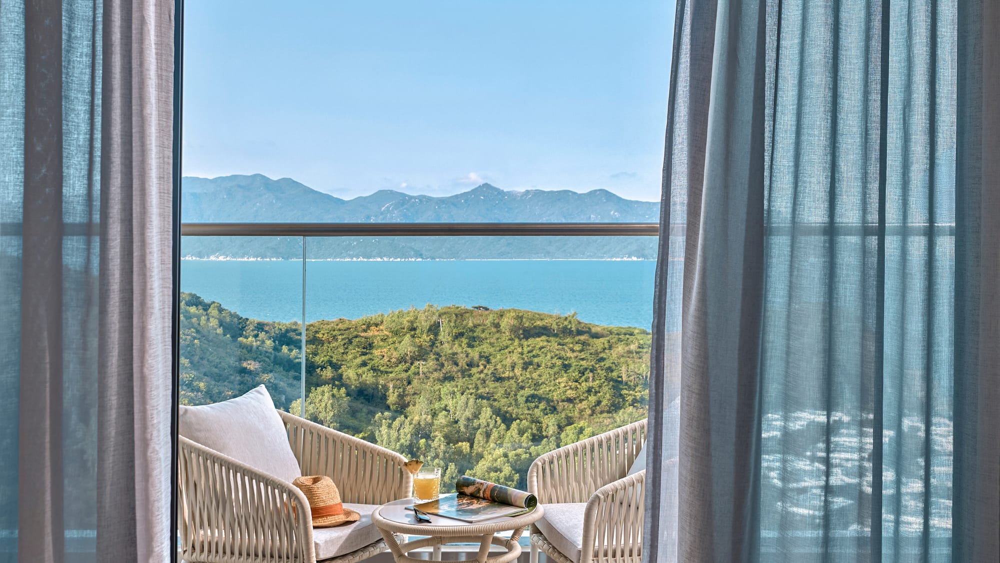 Balcony with sea view, outdoor seating, green hills, and blue sky.