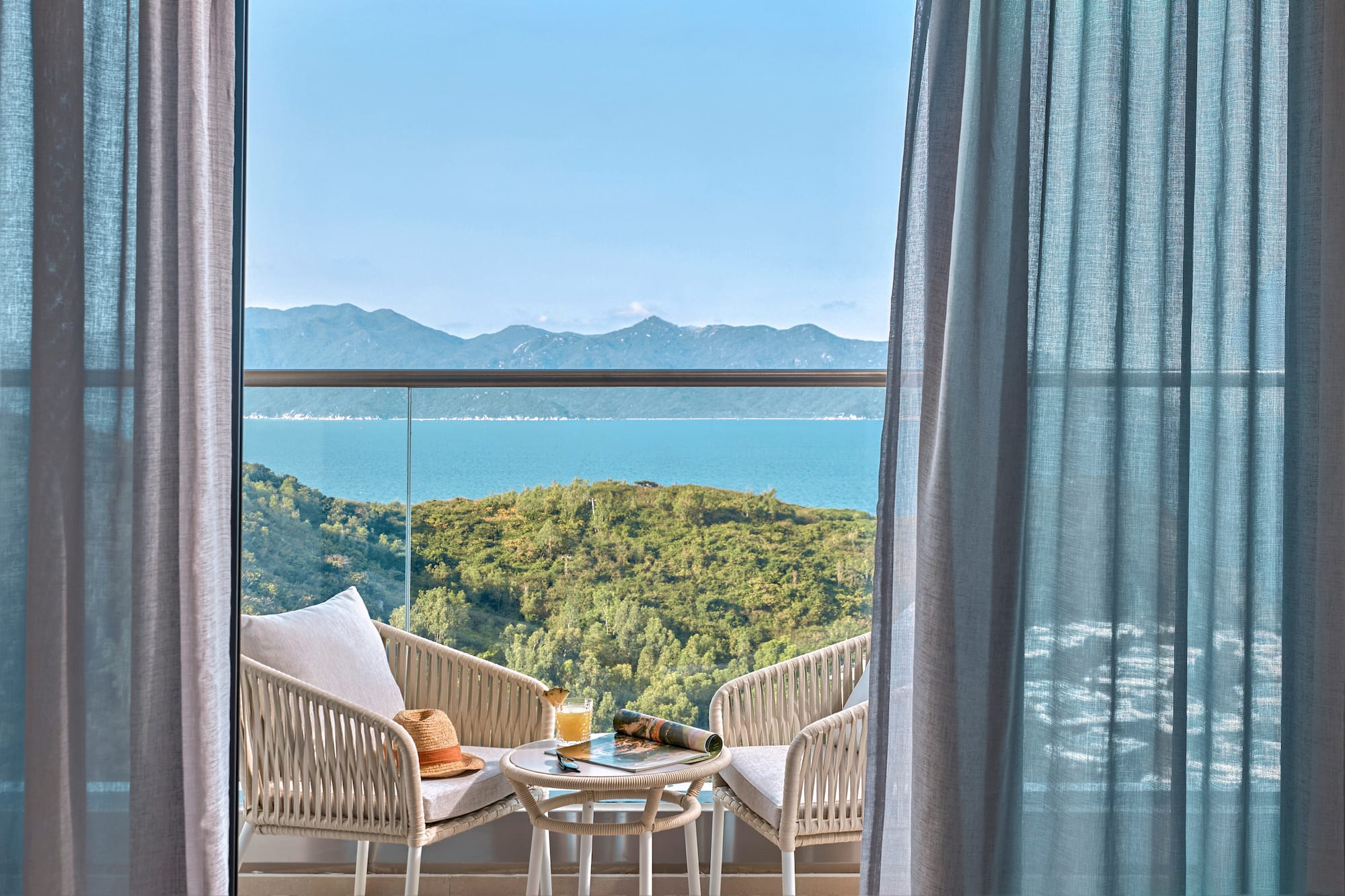 Balcony with sea view, outdoor seating, green hills, and blue sky.
