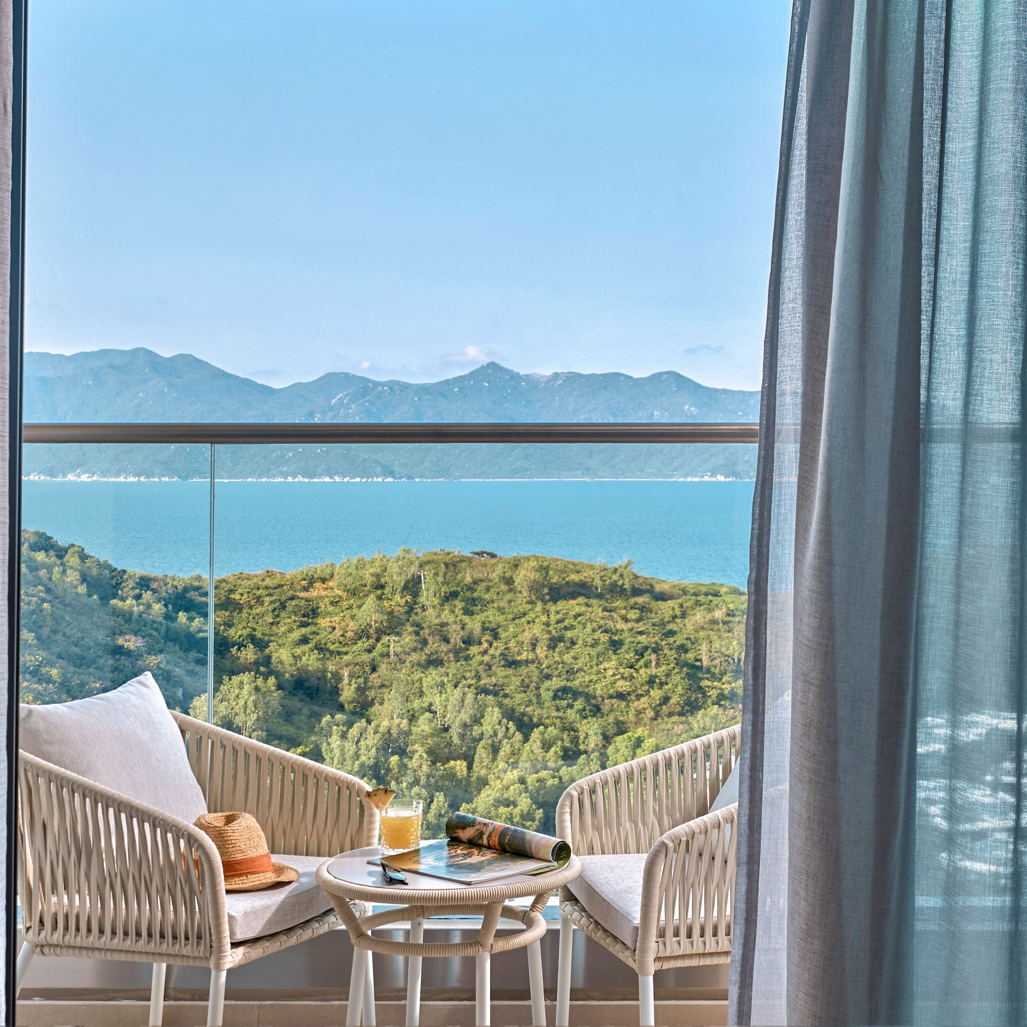 Balcony with sea view, outdoor seating, green hills, and blue sky.