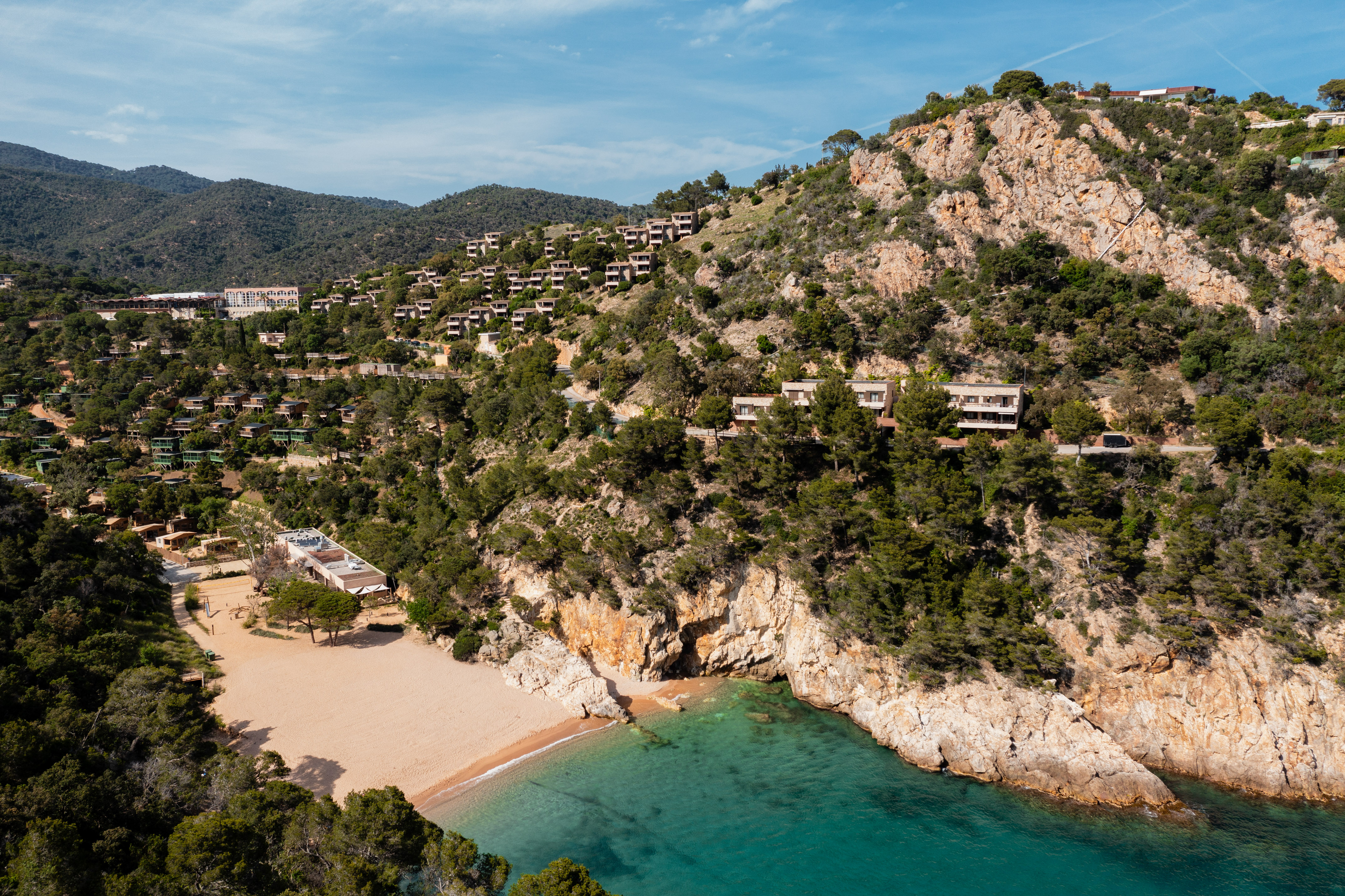 Secluded beach cove with turquoise water, rocky cliffs, and hillside resort.