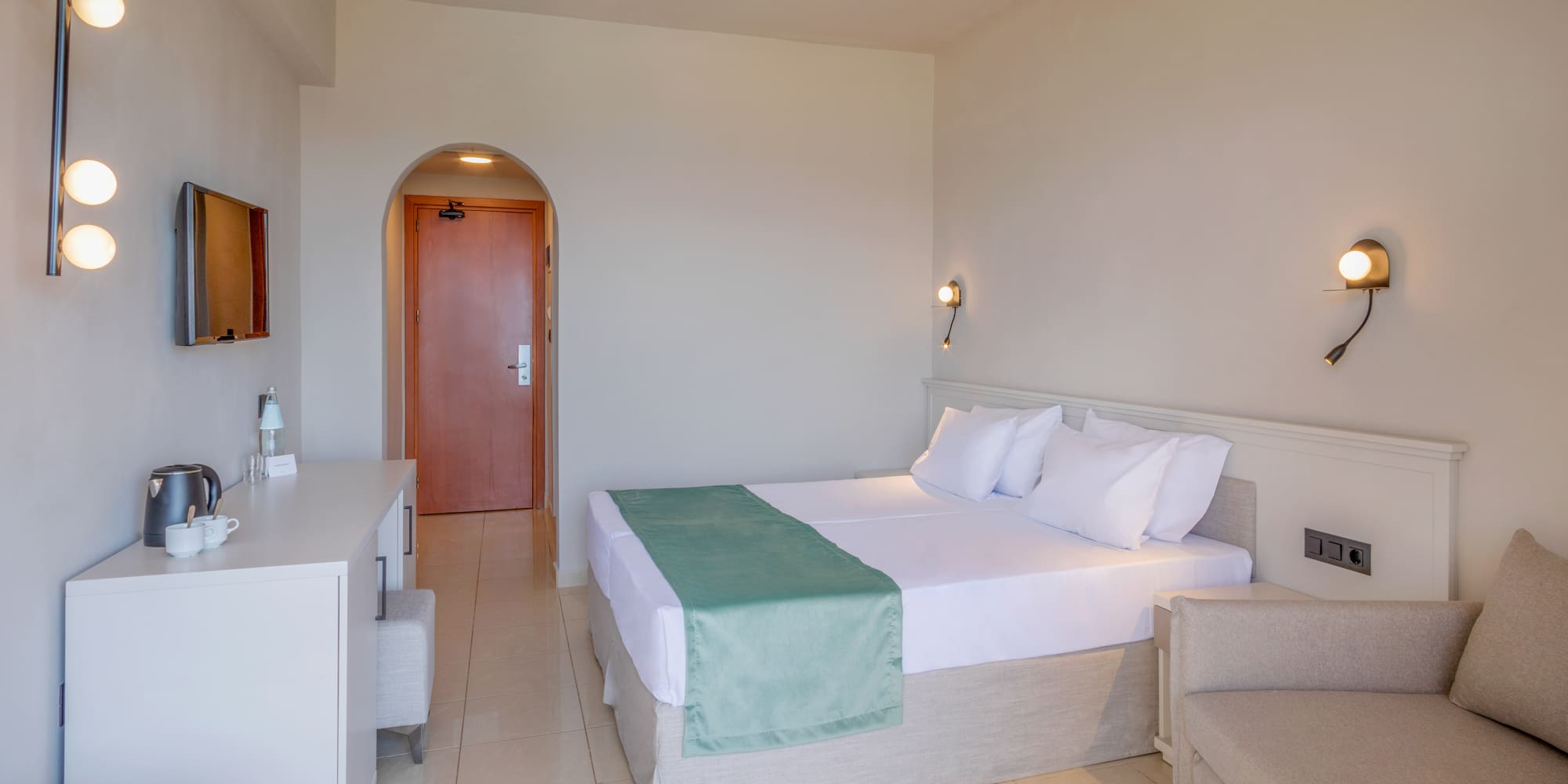 Modern hotel room with twin beds, green runner, TV, and lounge area.