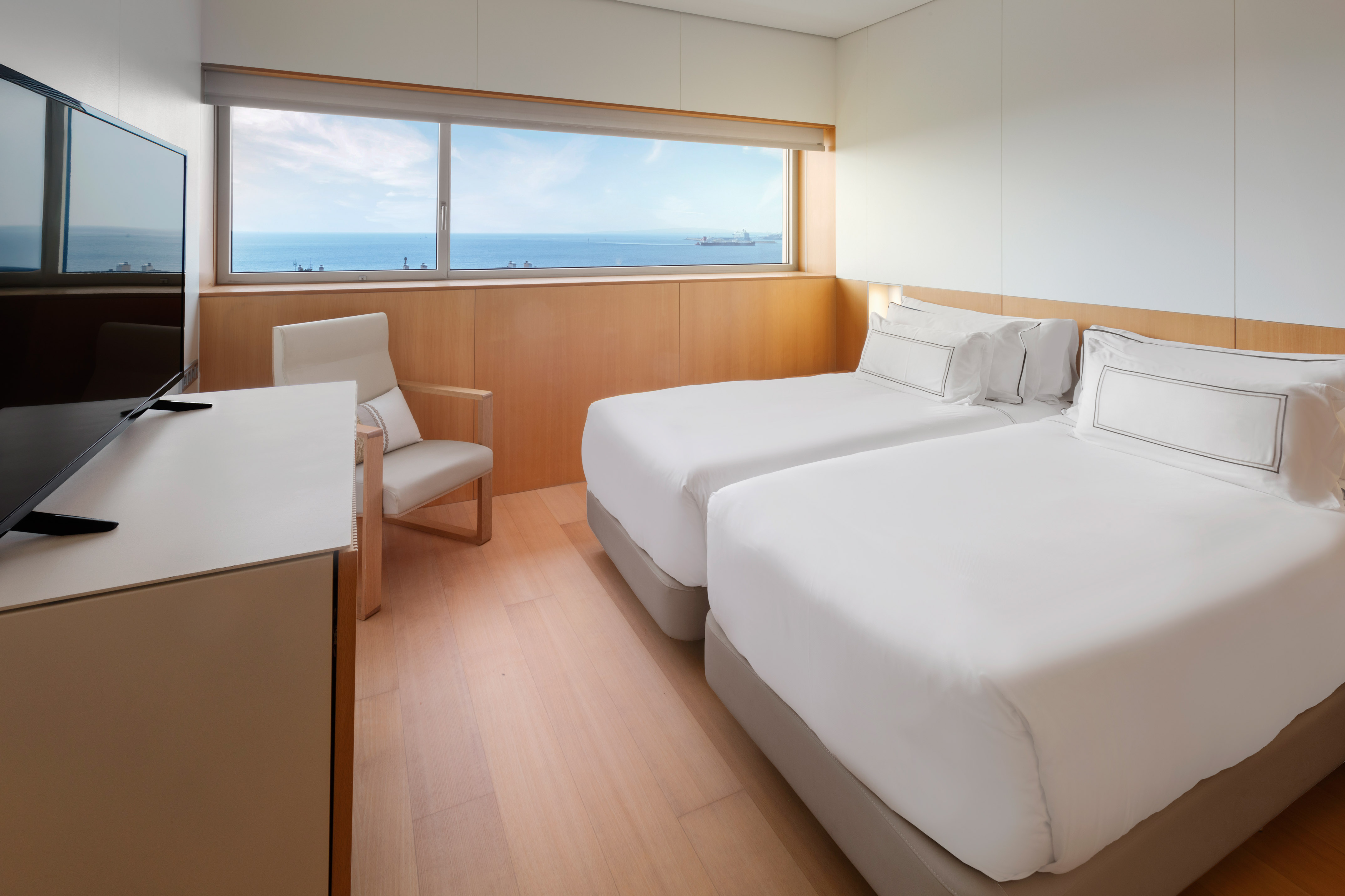 Bright modern hotel room with twin beds, ocean view window, and TV.