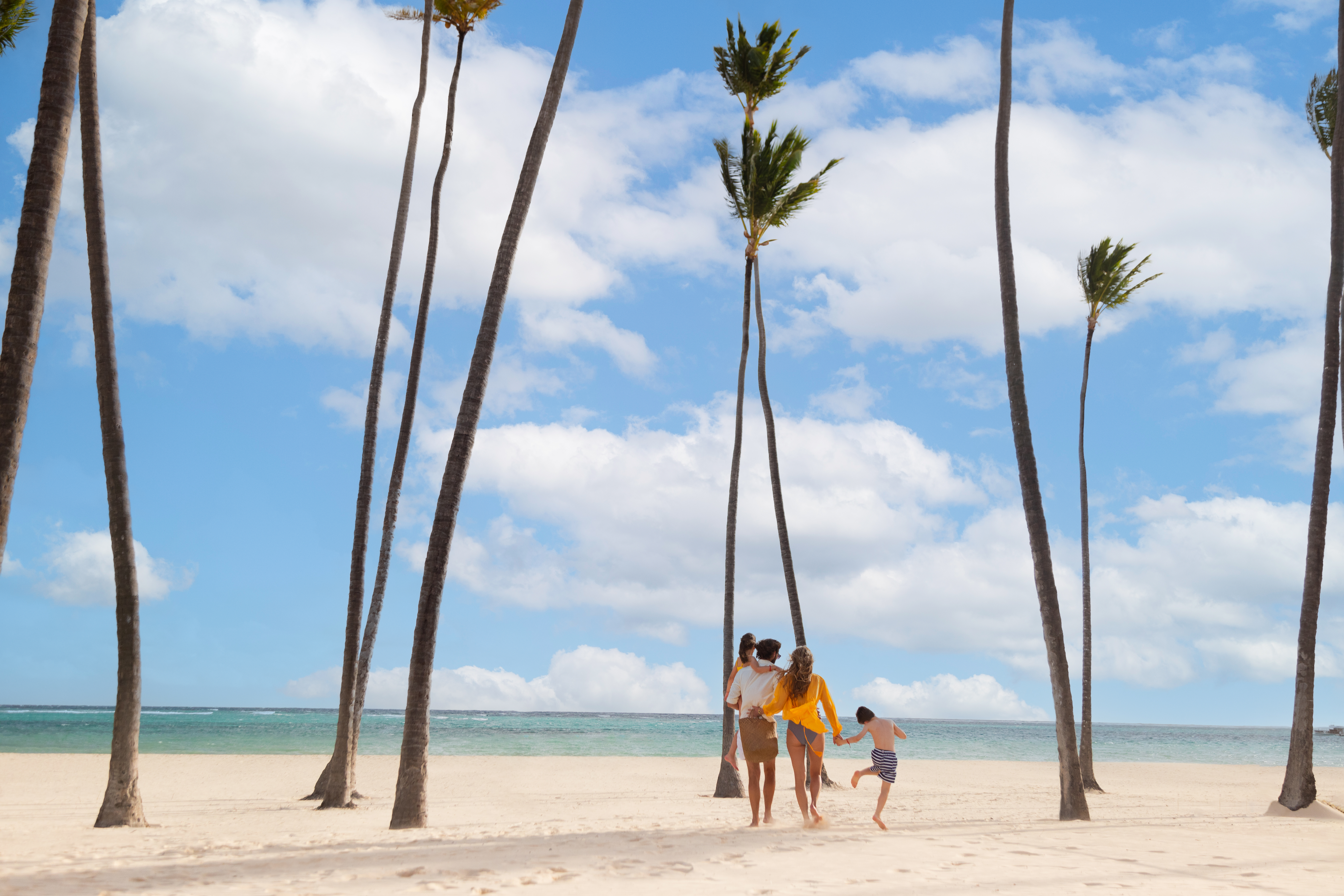 Family walks on tropical beach with palm trees, ocean, and blue sky.