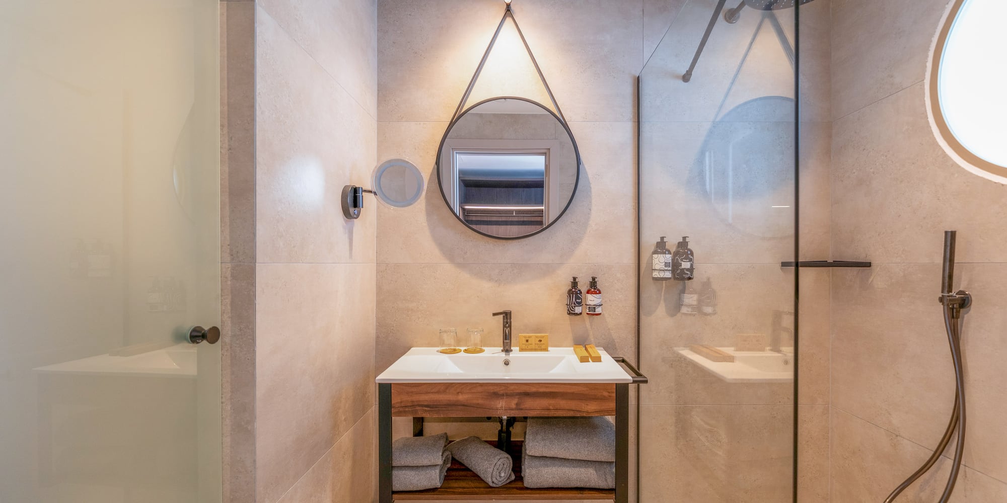 Hotel bathroom with modern wood vanity, round mirror, and glass shower.