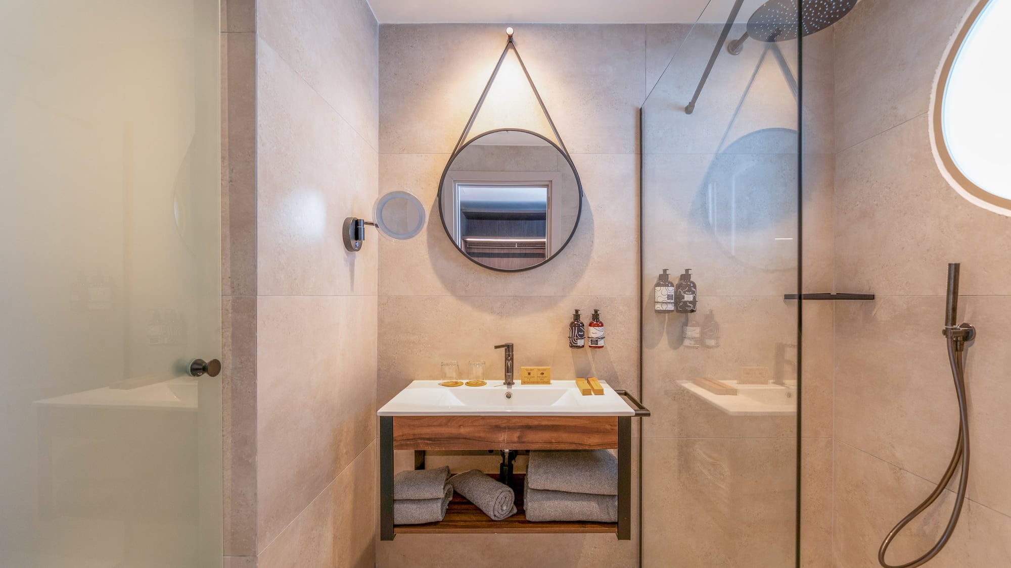 Hotel bathroom with modern wood vanity, round mirror, and glass shower.