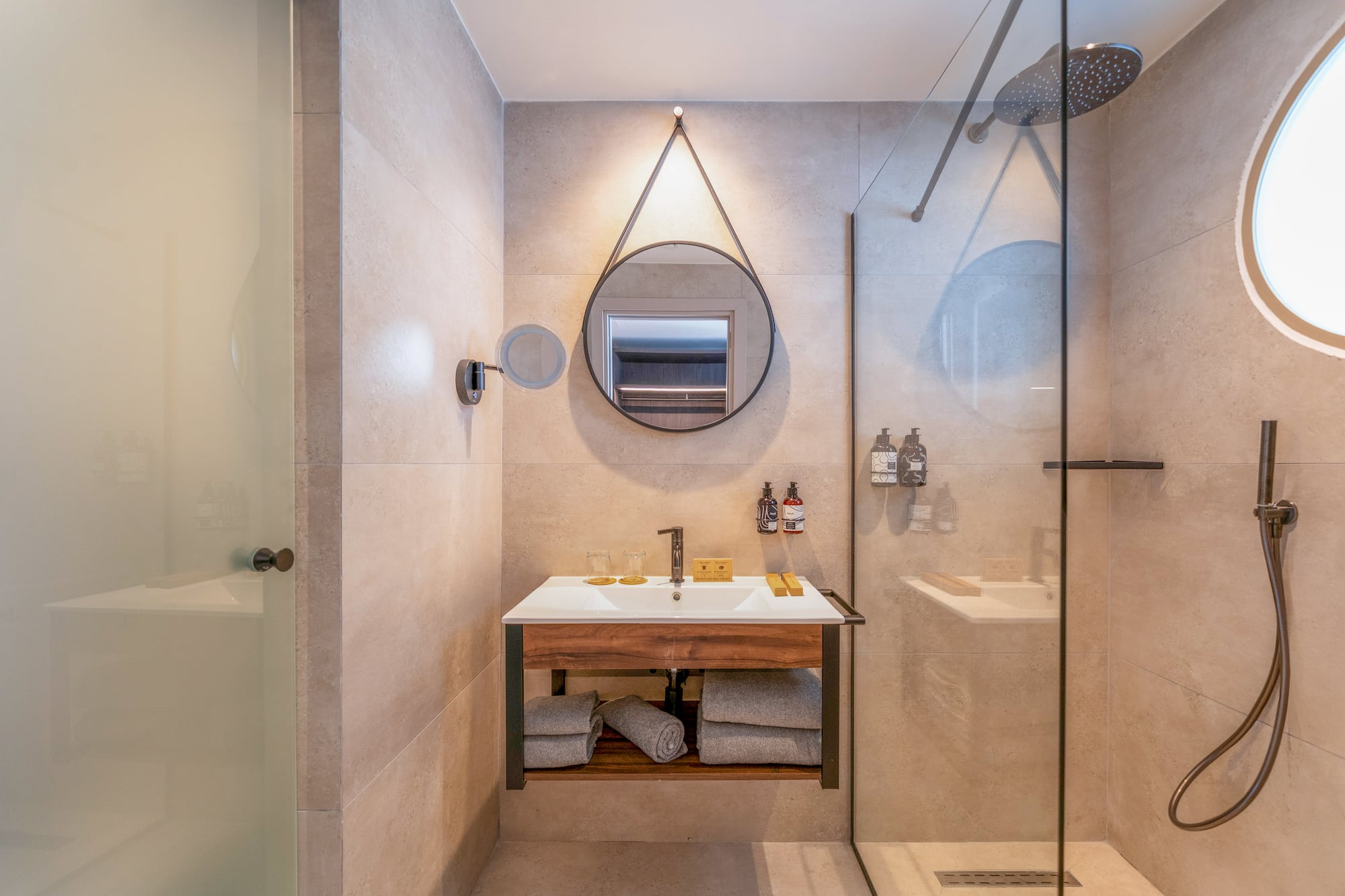 Hotel bathroom with modern wood vanity, round mirror, and glass shower.