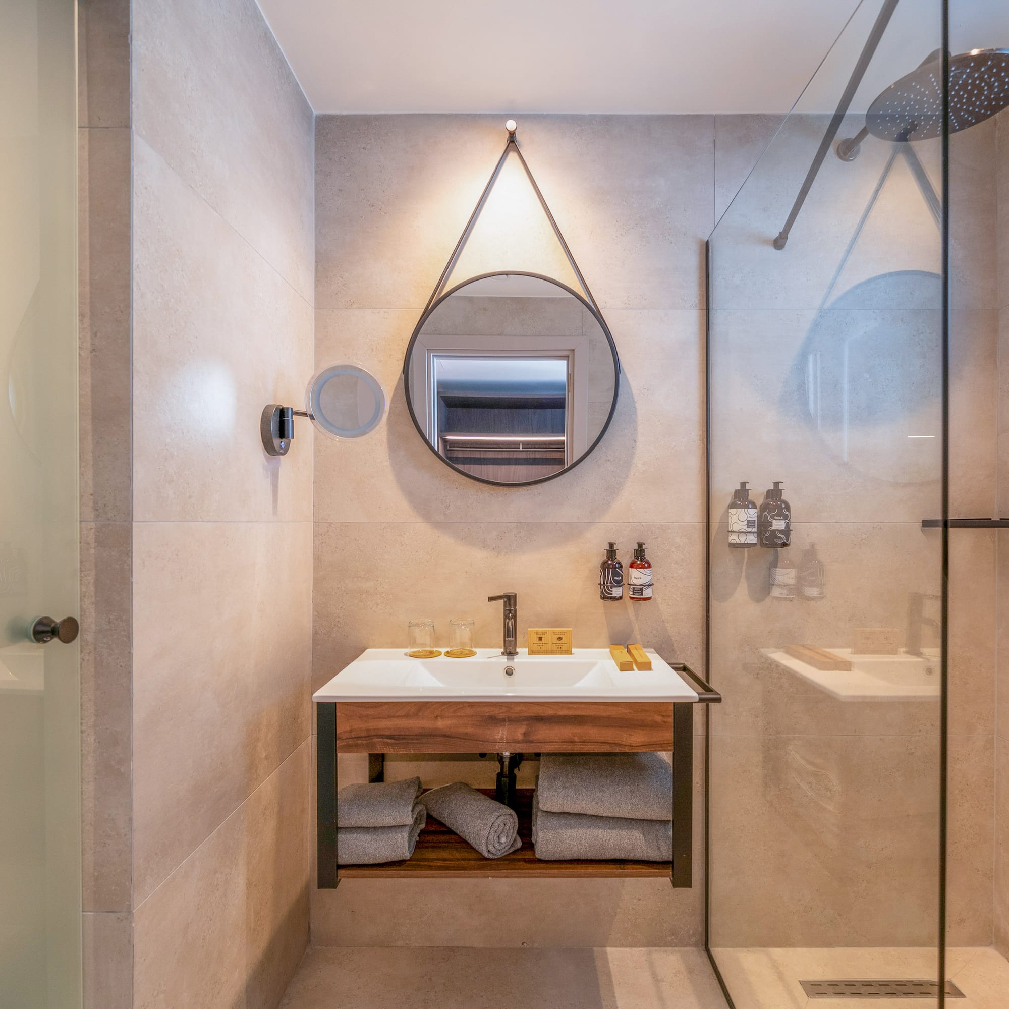 Hotel bathroom with modern wood vanity, round mirror, and glass shower.