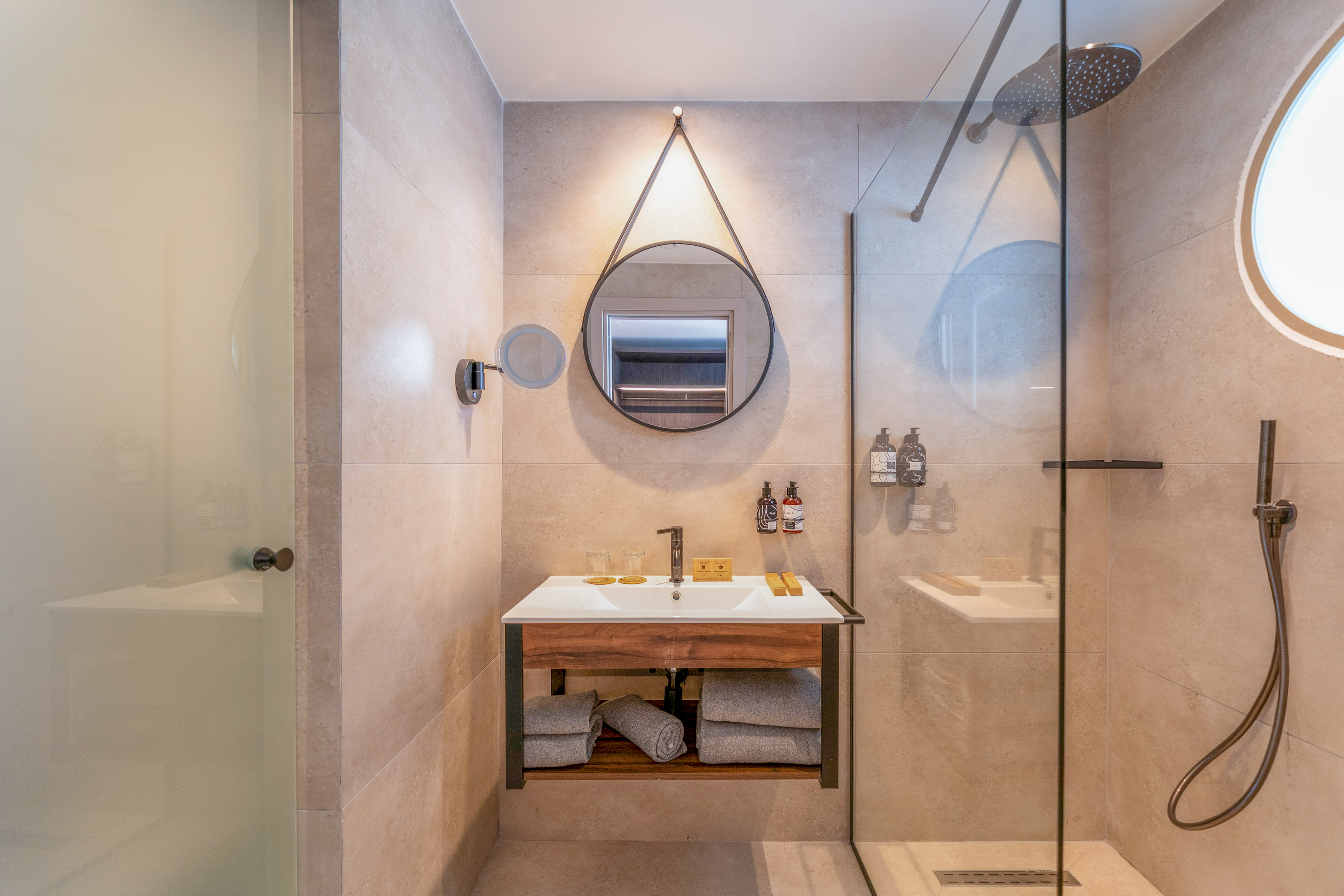 Hotel bathroom with modern wood vanity, round mirror, and glass shower.