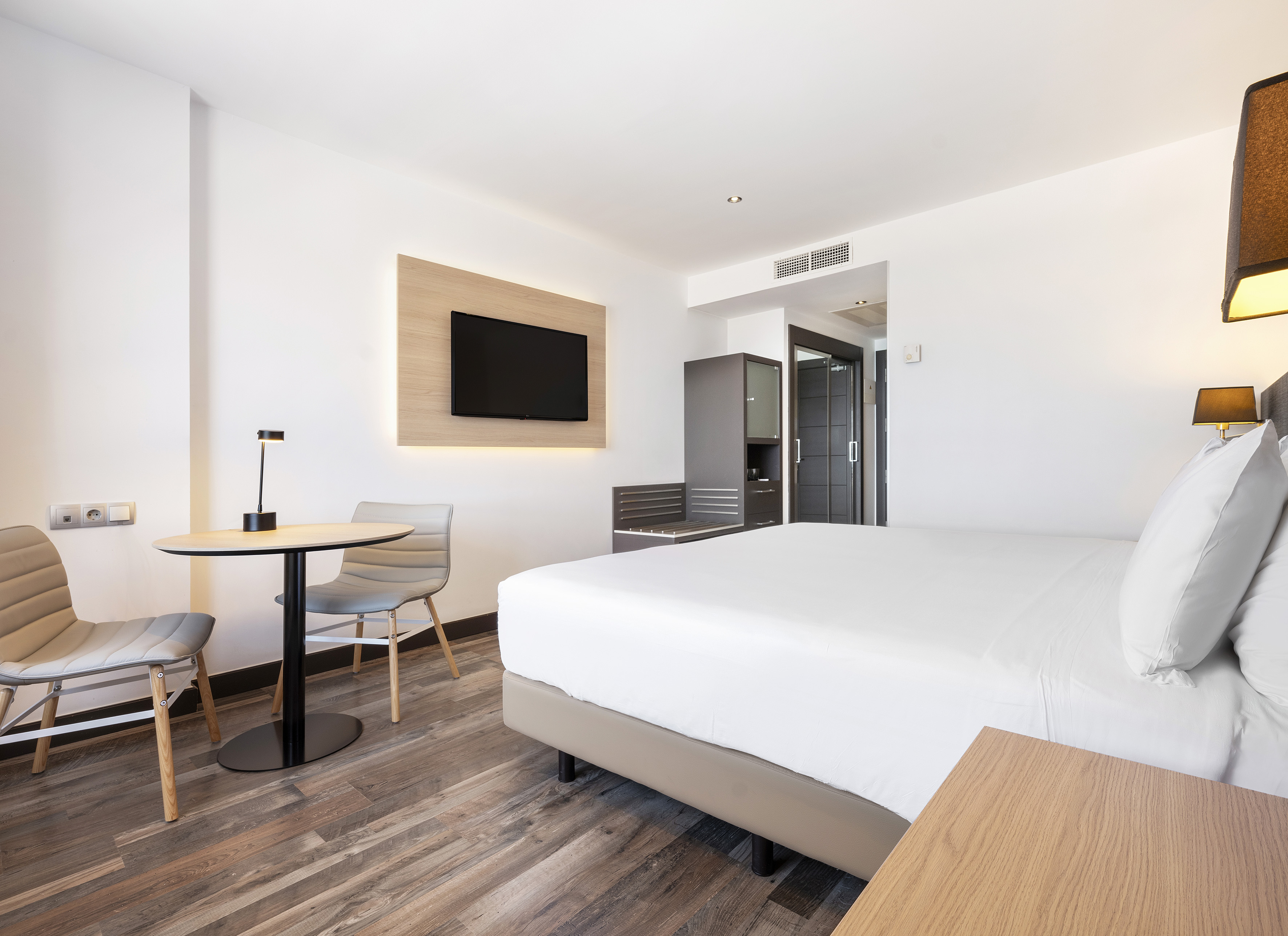 Modern guest room with white bed, wall-mounted TV, wood floor, and small table.