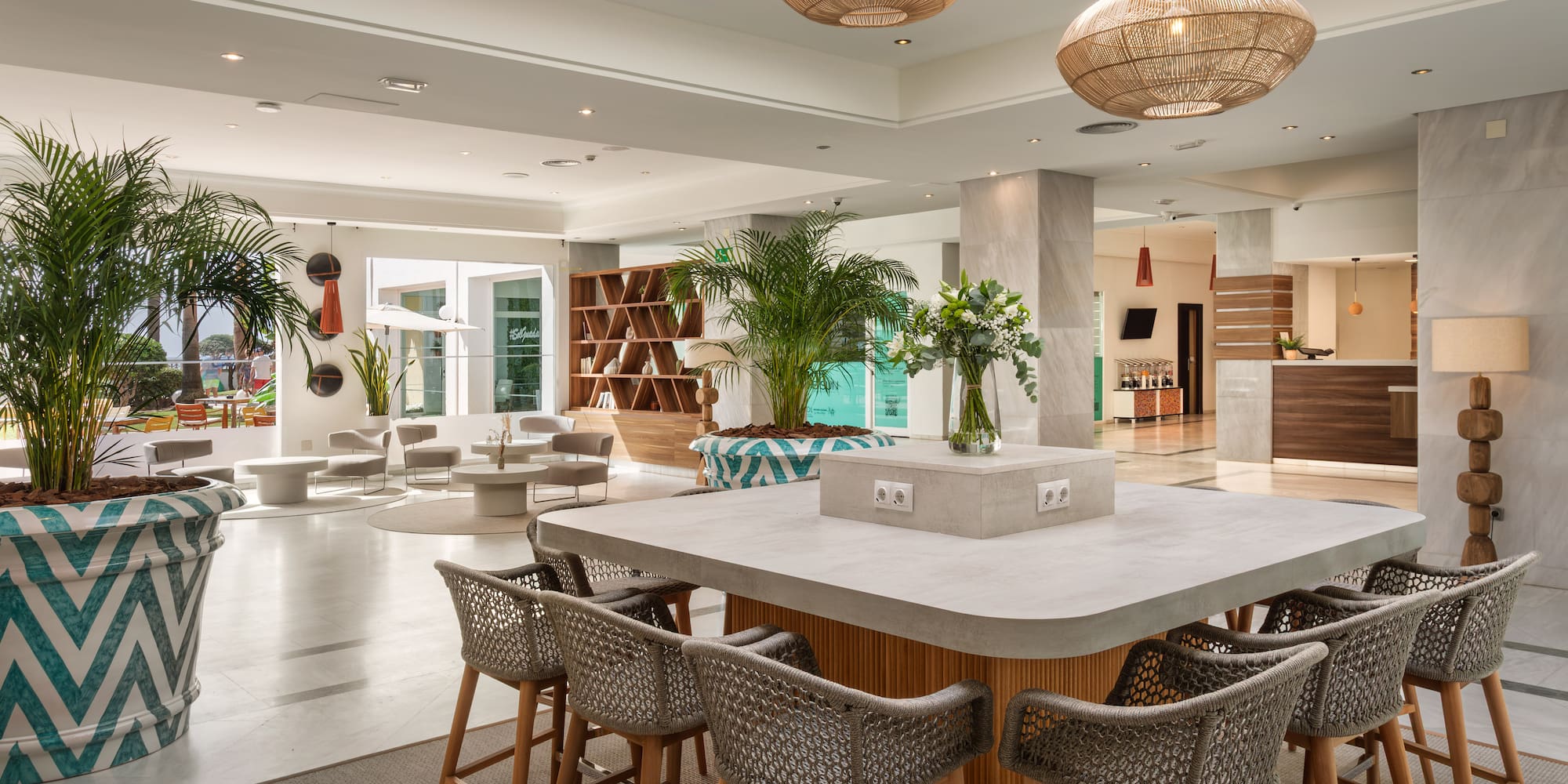 Modern hotel lobby lounge with central bar, tropical plants, and natural decor.