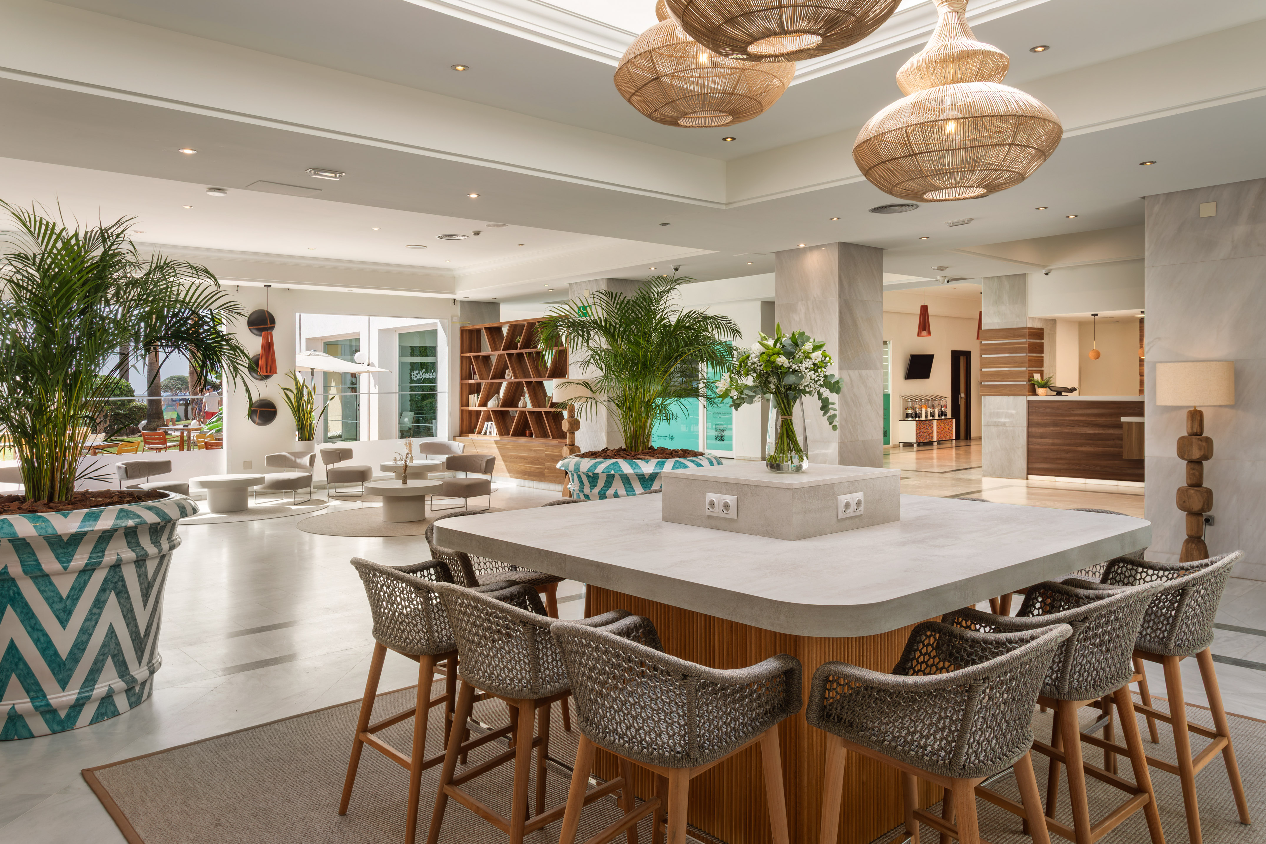 Modern hotel lobby lounge with central bar, tropical plants, and natural decor.