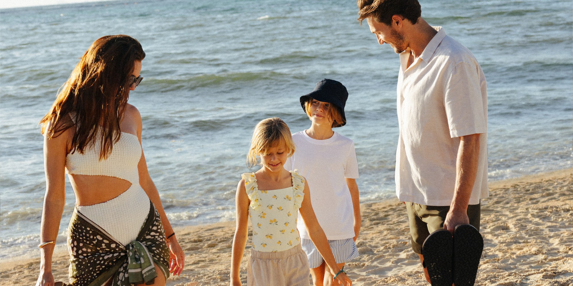 Family of four walking barefoot on a sunny beach by the ocean.