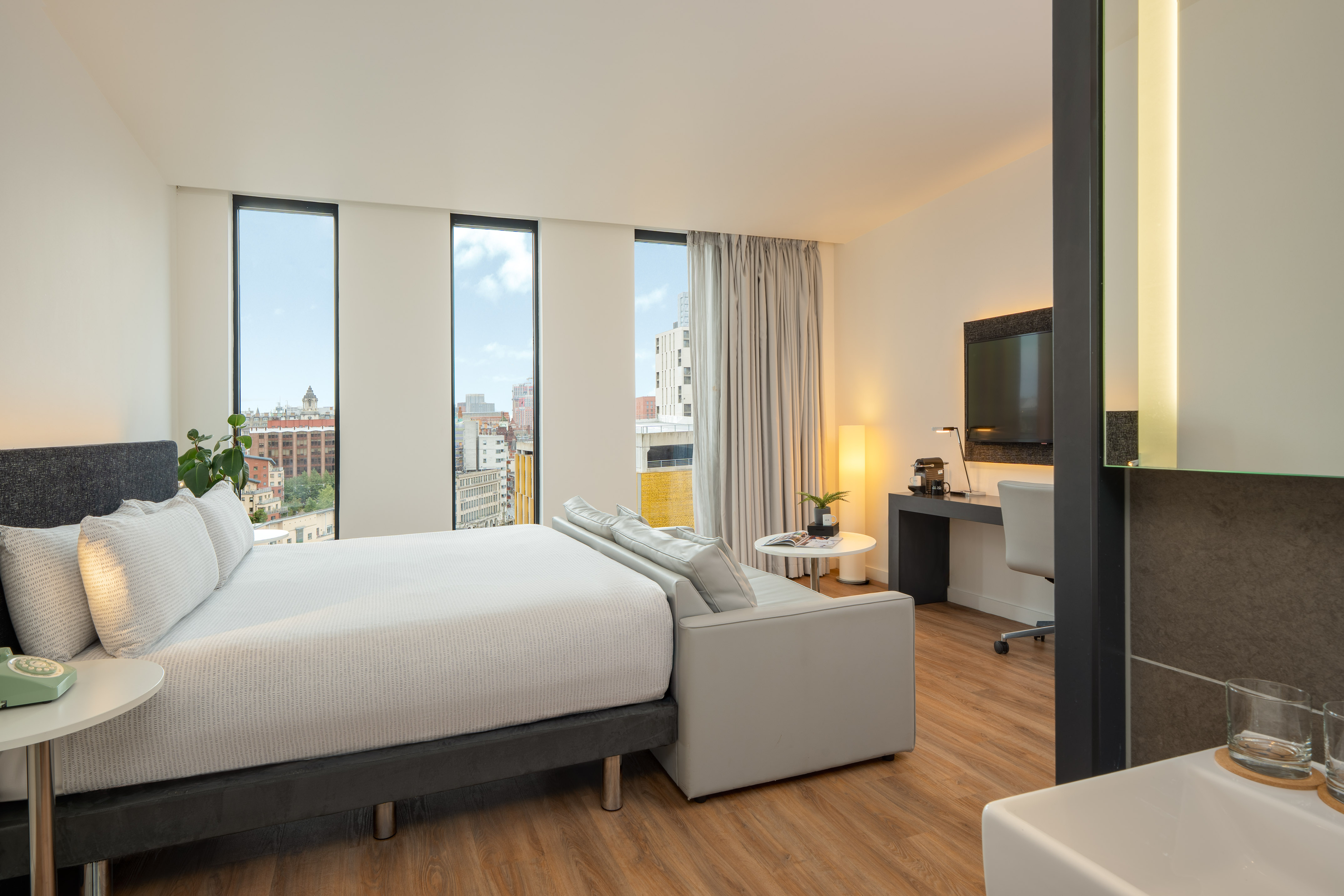 Modern hotel room with bed, sofa, desk, and city view windows.