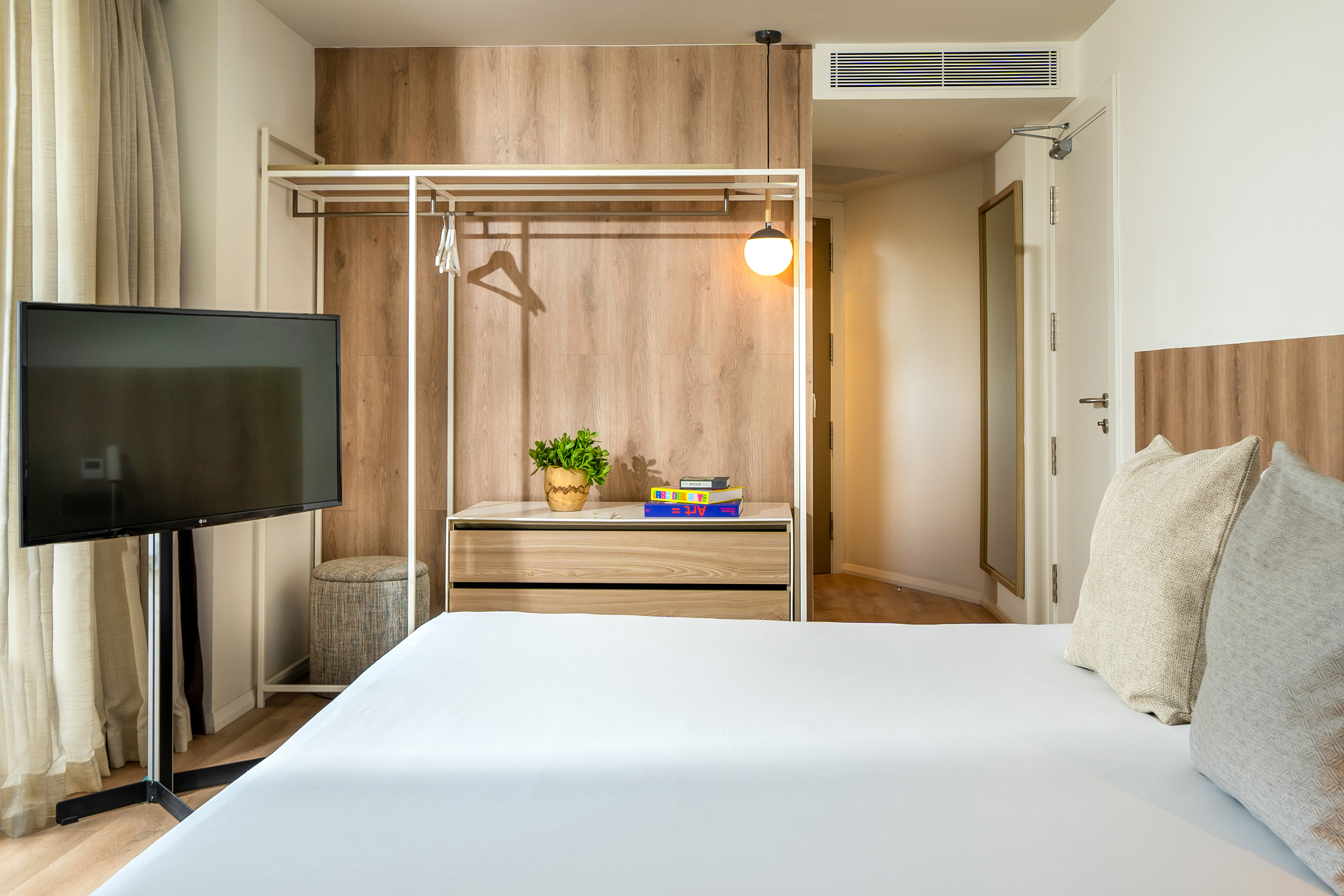 Modern hotel room with white bed, TV, open closet, and wood accents.