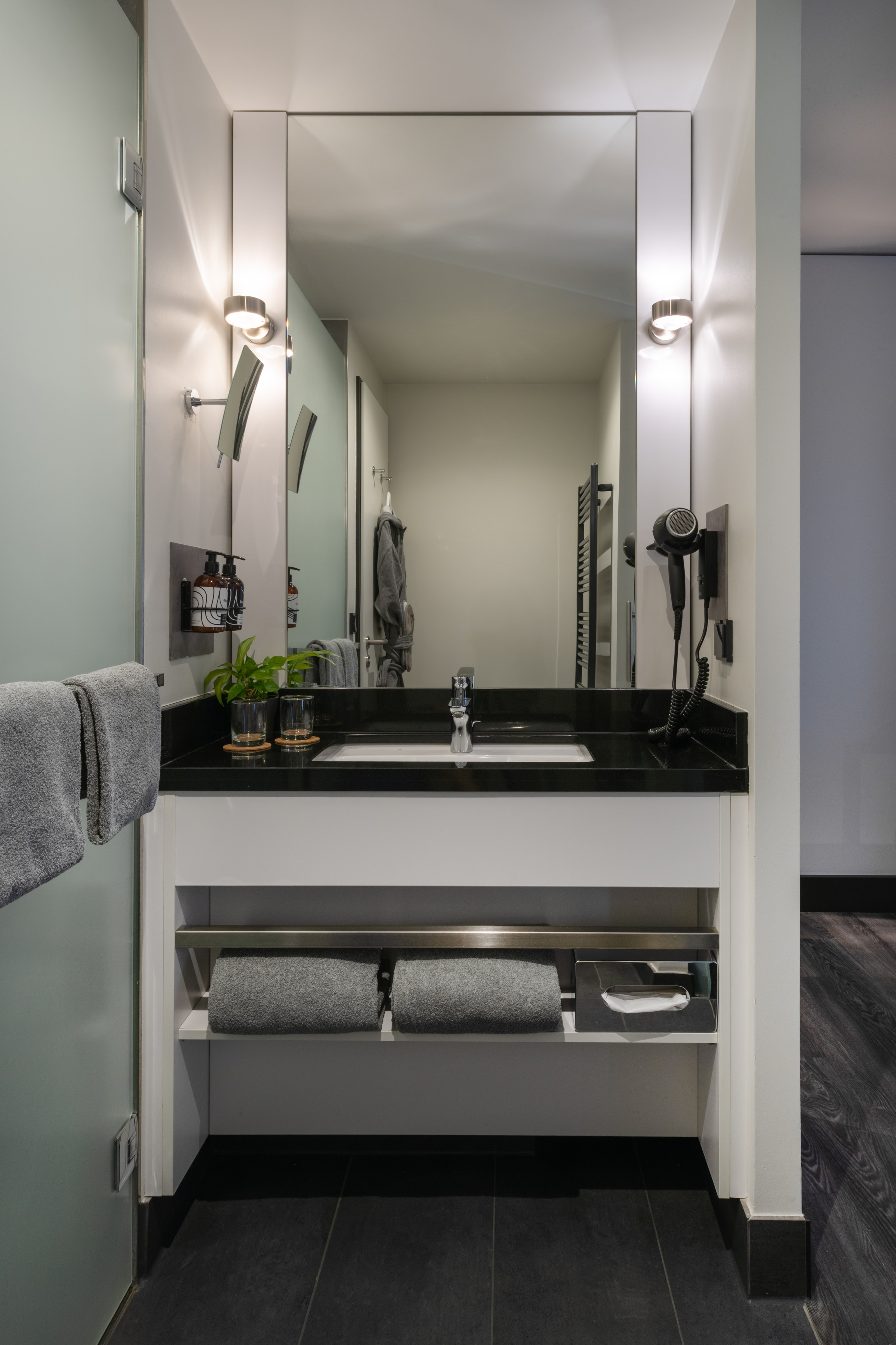 Modern hotel bathroom vanity with black counter, large mirror, sink, and grey towels. - The INNSiDE Room - INNSiDE Wolfsburg, Wolfsburg