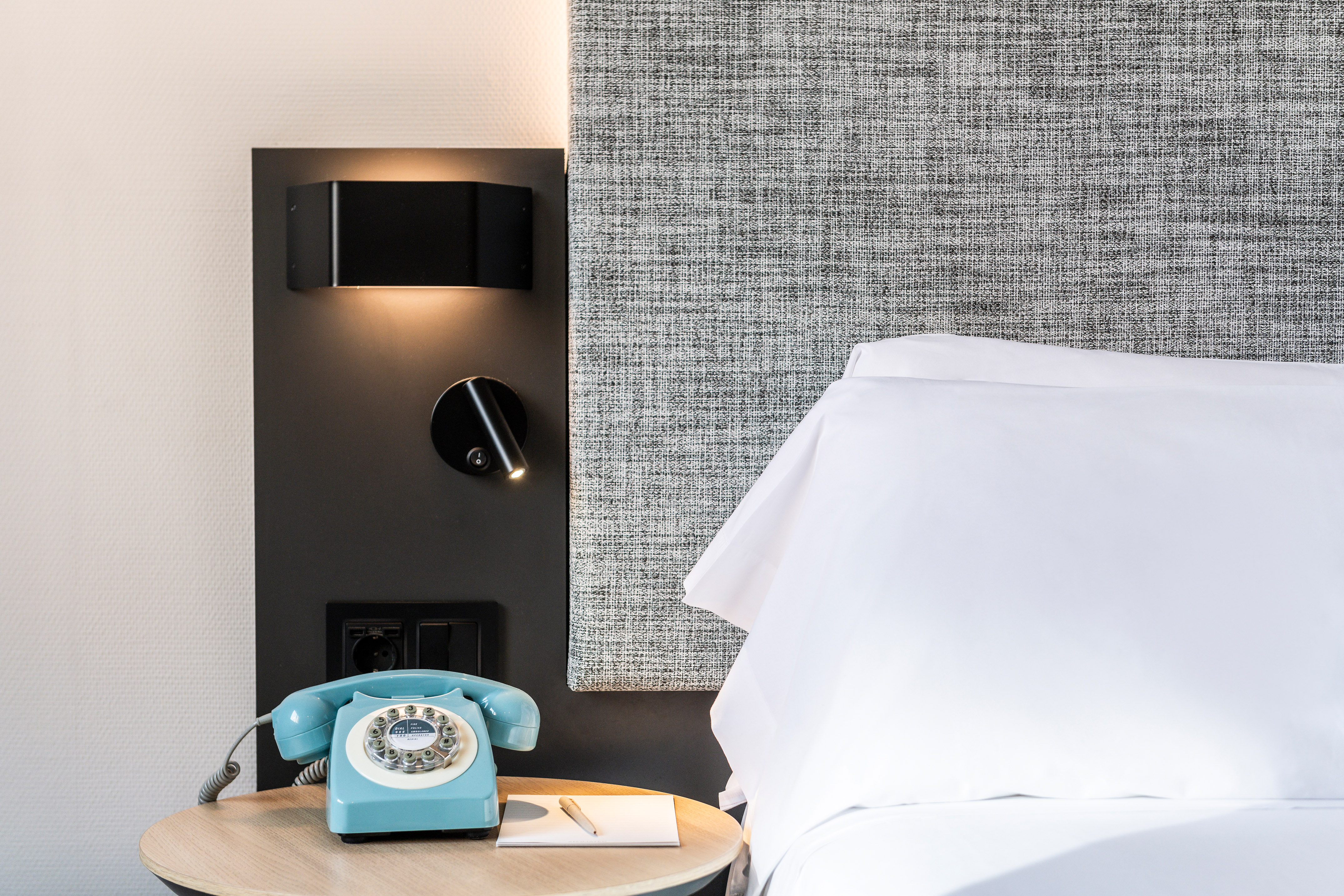 Modern hotel room with turquoise rotary phone, fabric headboard, and wall lamps.