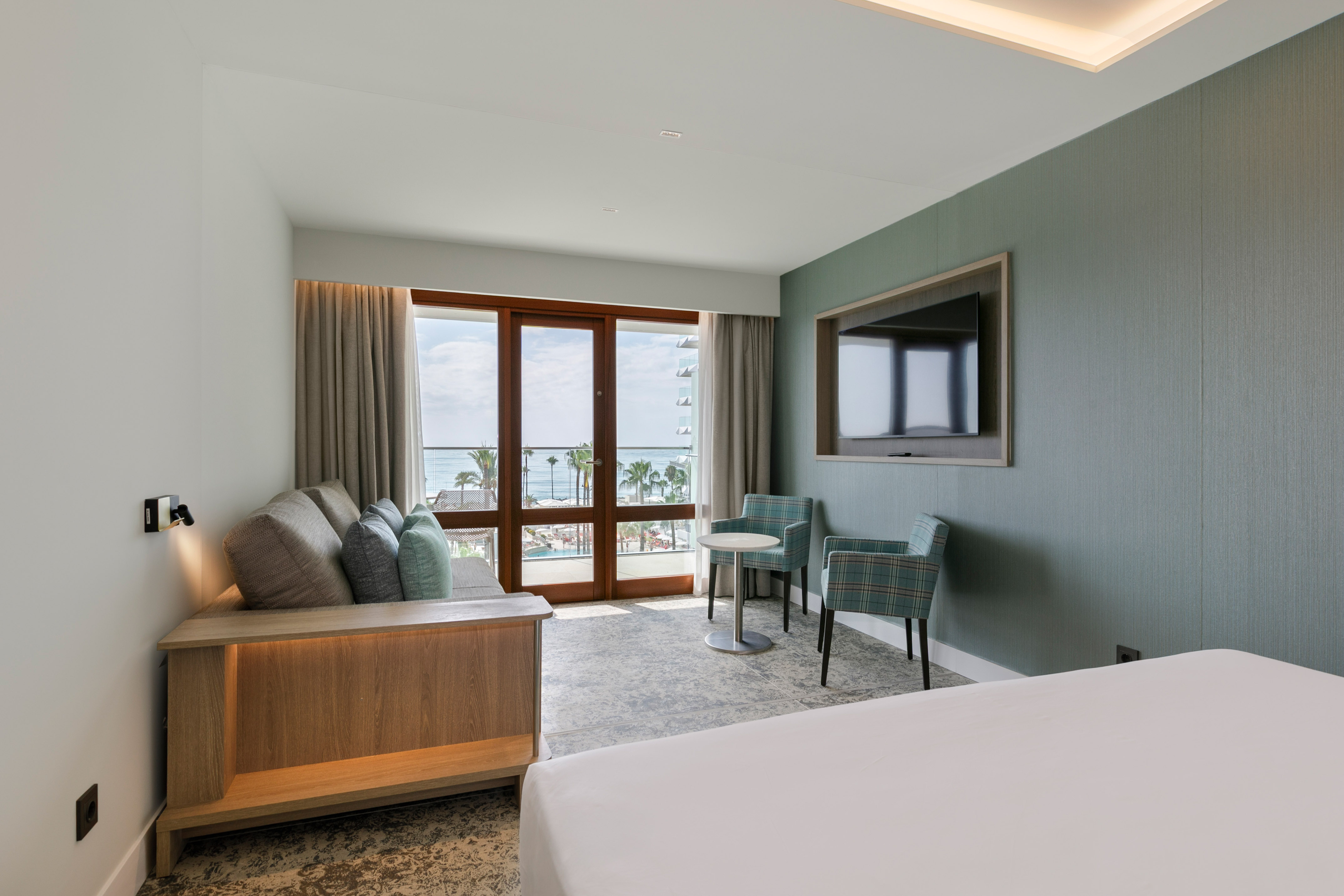Hotel suite with sea view balcony, sofa, armchairs, and wall TV.