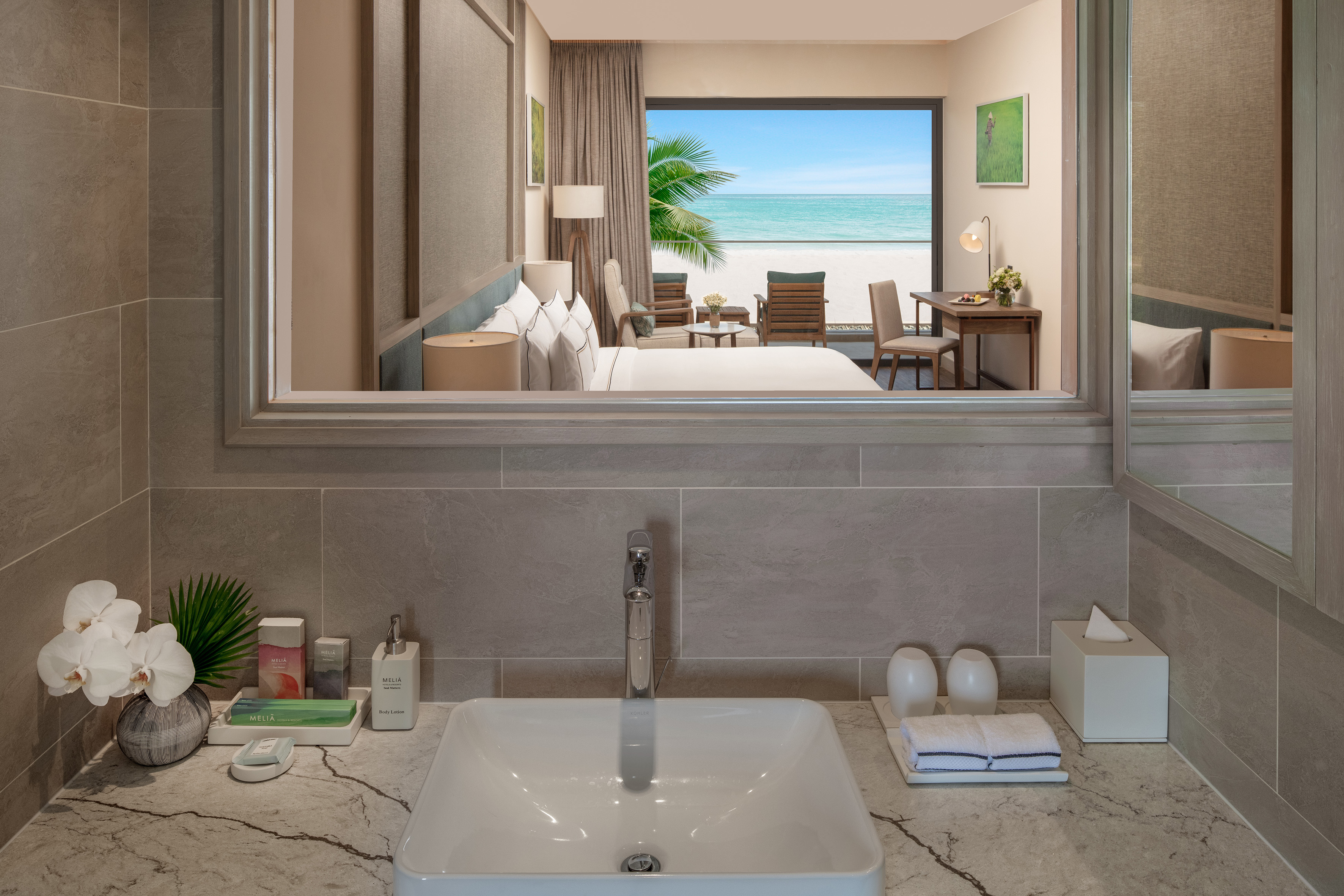 Hotel bathroom vanity reflecting bedroom, beach, and ocean view.