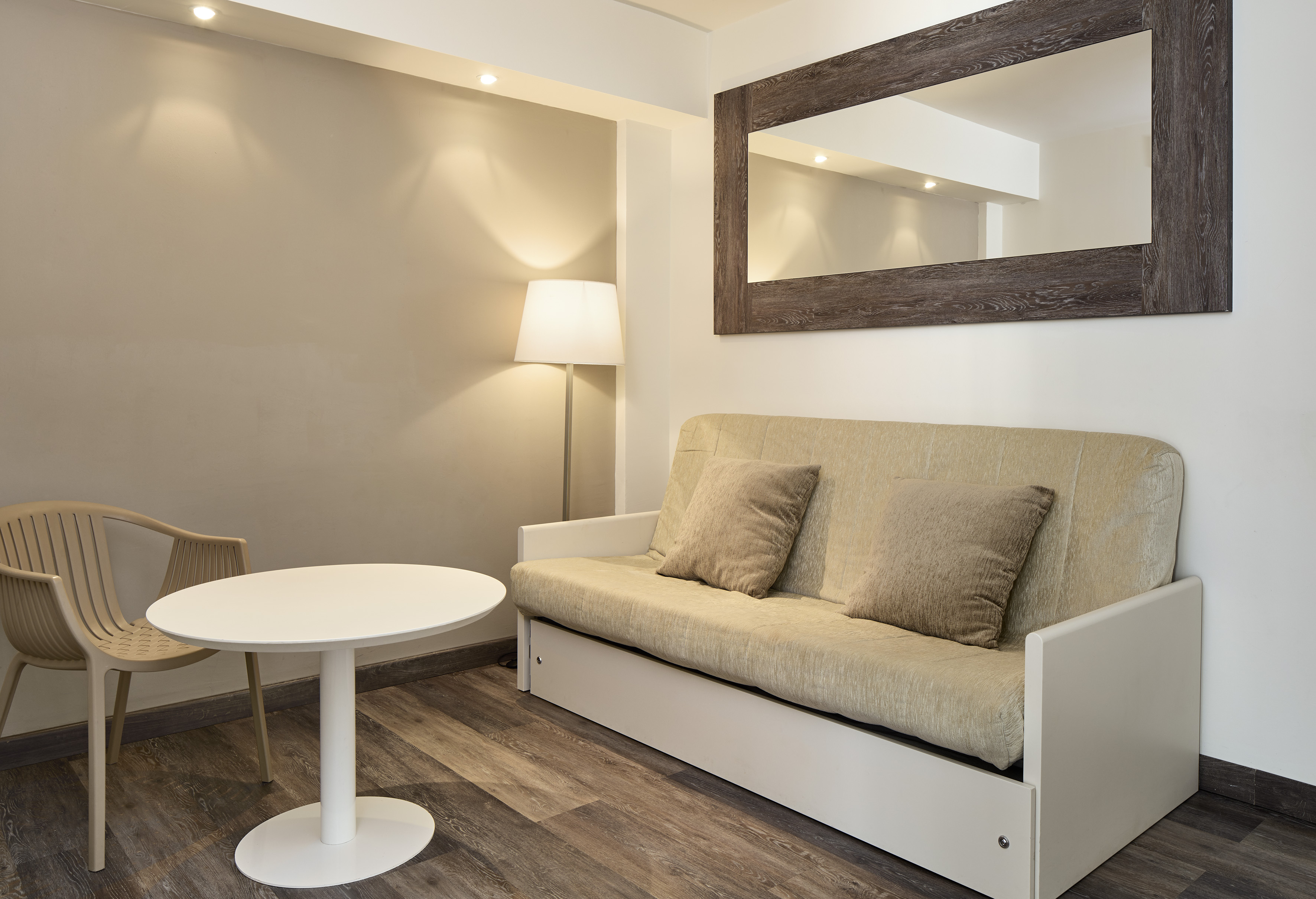 Neutral living space with beige sofa bed, white table, chair, lamp, and mirror.