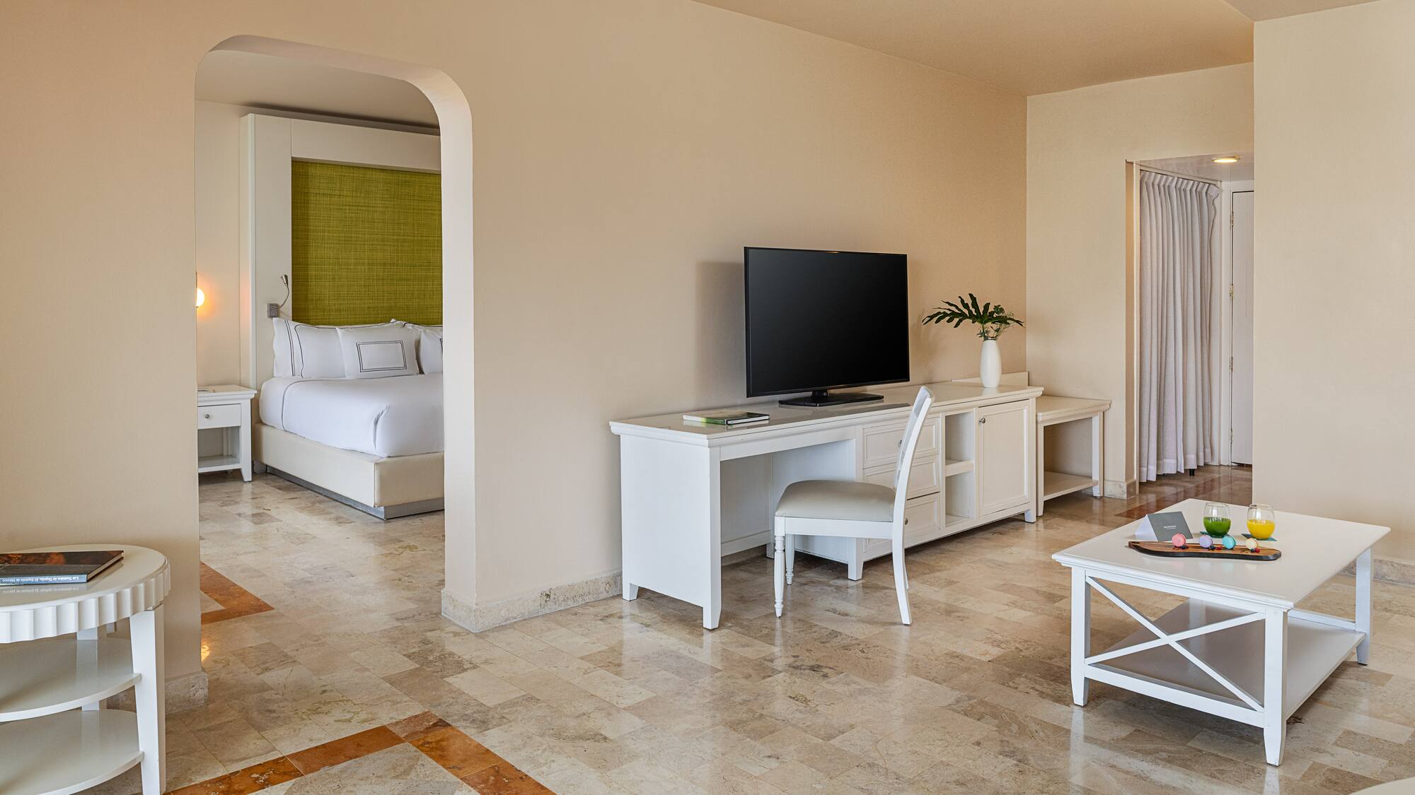 Hotel suite with connected bedroom, living space, TV, and white furniture.