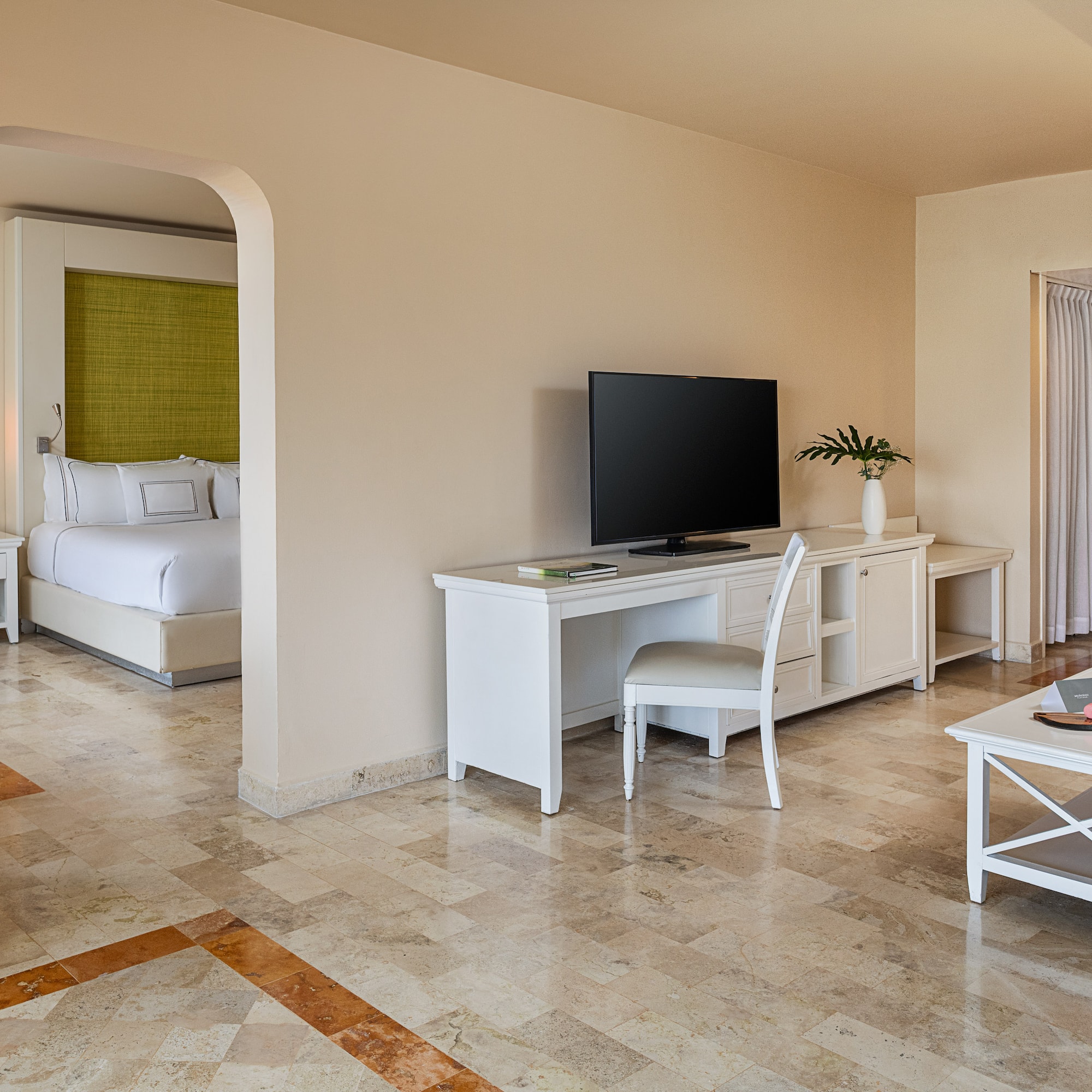 Hotel suite with connected bedroom, living space, TV, and white furniture.